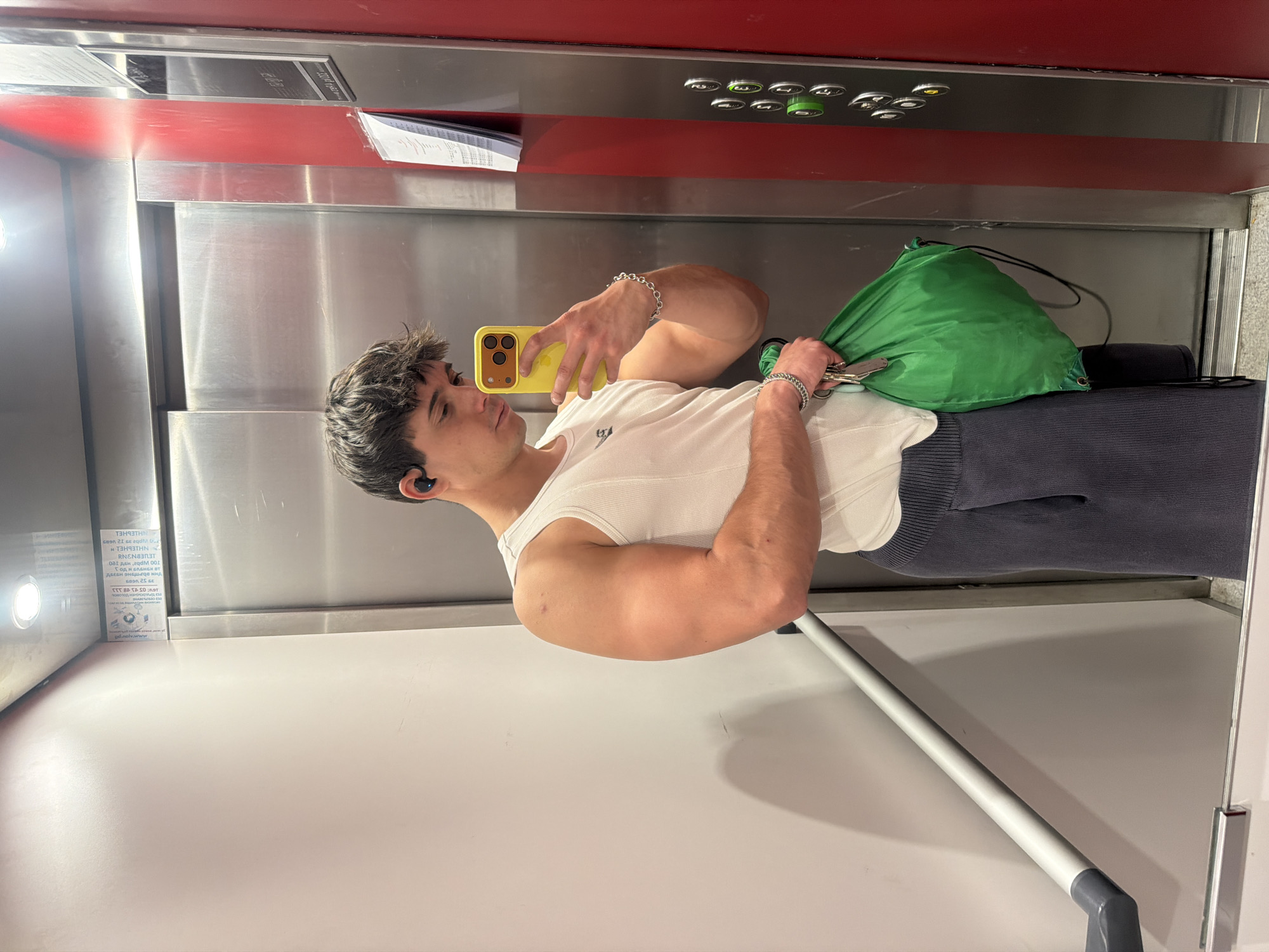The image shows a young man taking a mirror selfie inside an elevator. The man, with short, messy dark hair, is wearing a white ribbed tank top and dark, possibly navy, sweatpants. He holds a yellow smartphone with three cameras in his right hand, looking at the screen as he takes the photo. A silver chain bracelet is visible on his left wrist, and a black earbud is in his left ear. In his left hand, he holds a bunch of keys and a bright green drawstring bag.
The elevator interior features metallic walls and a red panel above the control panel. The control panel has several circular buttons, some appearing green. A light-colored handrail is visible in the lower right. On the left metallic wall, a mirrored sign with Cyrillic text is reflected, revealing words like "ИНТЕРНЕТ" (Internet), data capacities such as "25 GB," "60 GB," "100 GB," and a phone number starting "T: 02 88-88-88." This text suggests the location is Sofia, Bulgaria, consistent with the provided context. The man's expression is neutral and focused on taking the picture.