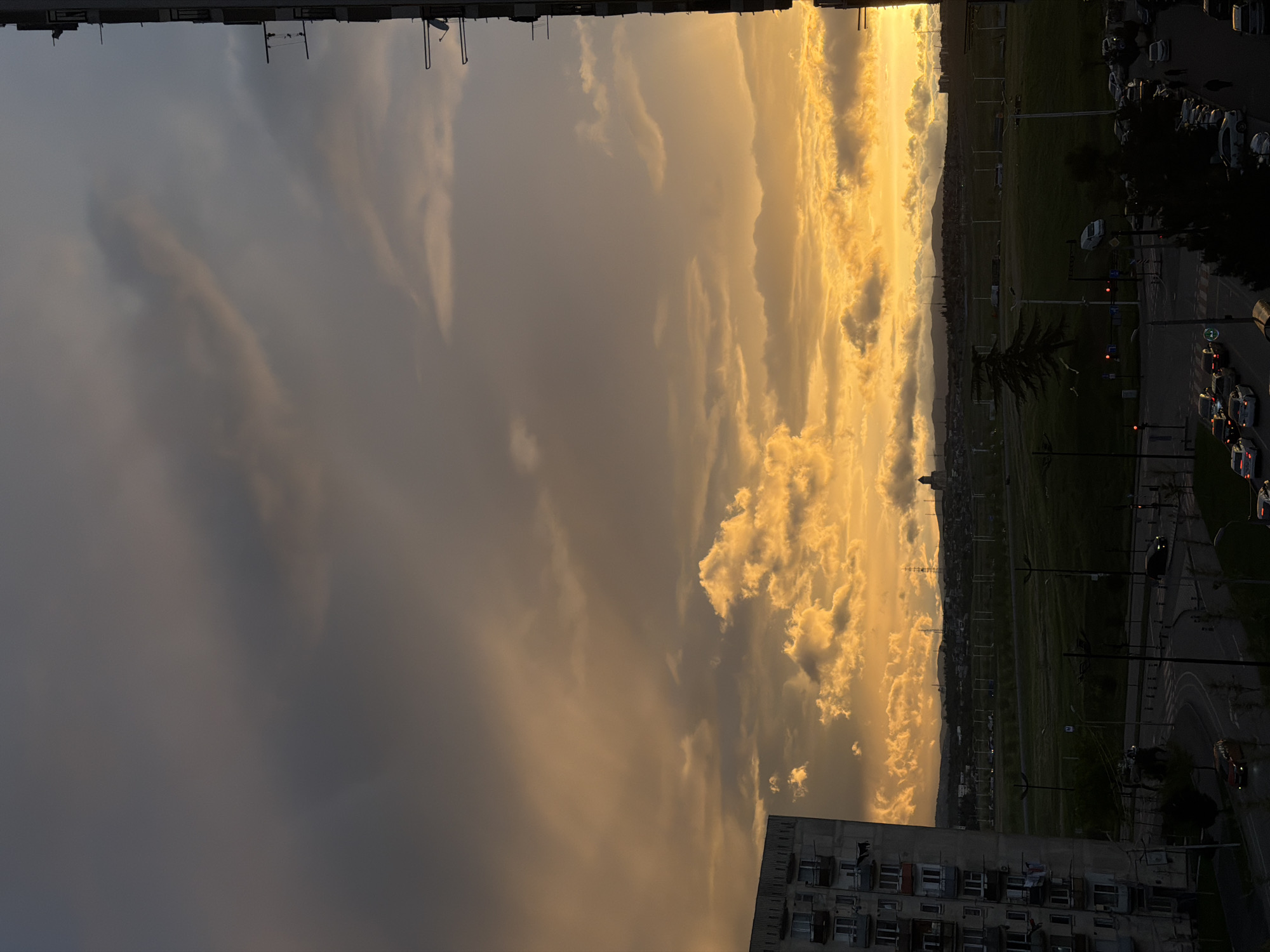 The image presents a view of a city, identified as Tbilisi, Georgia, during what appears to be either sunrise or sunset. The sky dominates the scene, with a dramatic display of clouds colored in gradients from dark gray to bright orange and yellow, suggesting the presence of the sun near the horizon.
Below the sky, a landscape consisting of buildings, streets, and green spaces is visible. The urban environment includes a road with moving cars and parked vehicles. The buildings appear to be a mix of residential and commercial structures. The overall environment suggests a residential area of Tbilisi.