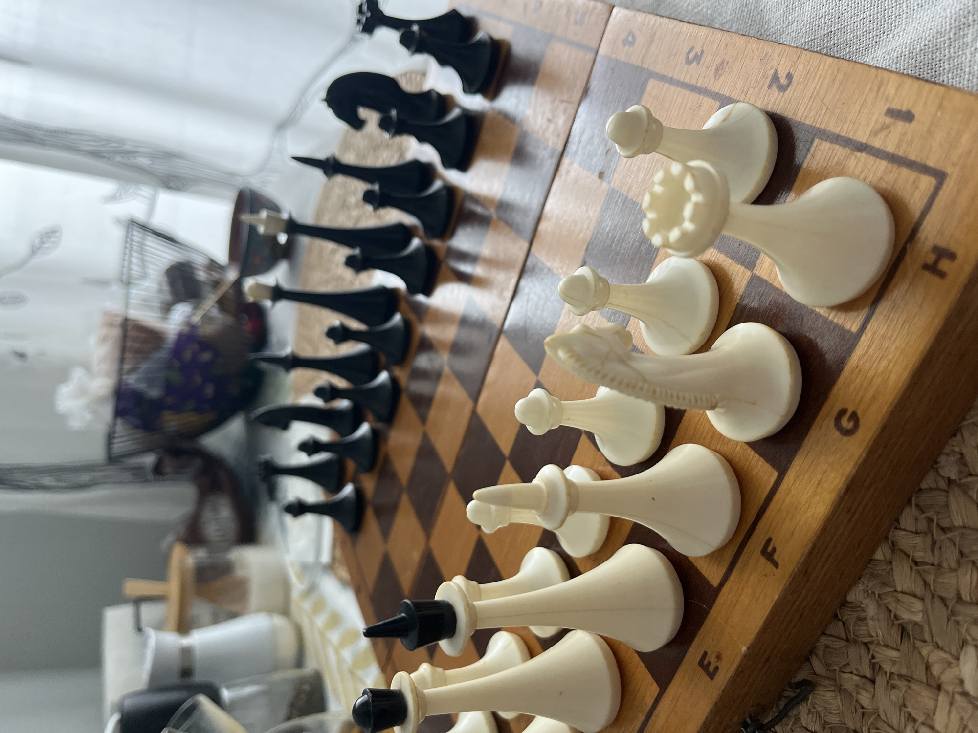 A chess set with black and white pieces is arranged on a wooden chessboard. The board has a brown and tan square pattern. The white pieces are closer to the foreground, and the black pieces are farther away. Letters (A-H) and numbers (1-8) are etched around the edge of the board. The chessboard is sitting on a textured, possibly woven, surface. There is a blurry background, including a white curtain and a wire basket.
There is no information regarding the city of Izhevsk, Russia.