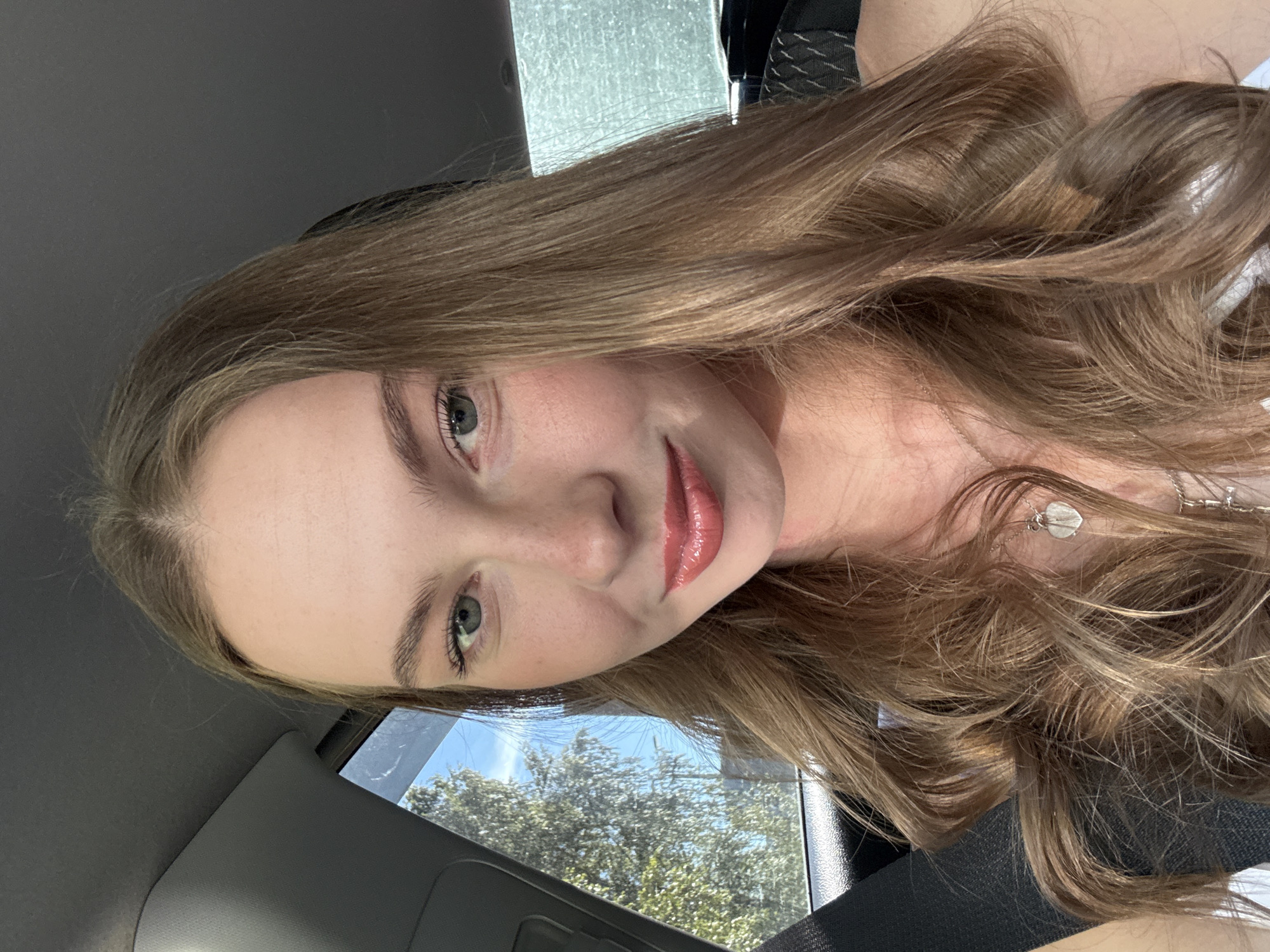 This is a portrait-oriented, low-angle selfie of a young woman with long, wavy, light brown hair. She is wearing a light-colored top and a silver necklace with a heart-shaped pendant. The background appears to be the interior of a car, with a window revealing a glimpse of trees and a bright, sunny sky. The lighting suggests it is daytime. The woman is looking towards the camera with a gentle smile and her lips are coated with a shiny lip gloss. Her eyes are blue-green and she has natural-looking makeup. The image is well-lit and the focus is on the woman's face and hair. There is no visible text in the image. The overall mood is casual and relaxed.