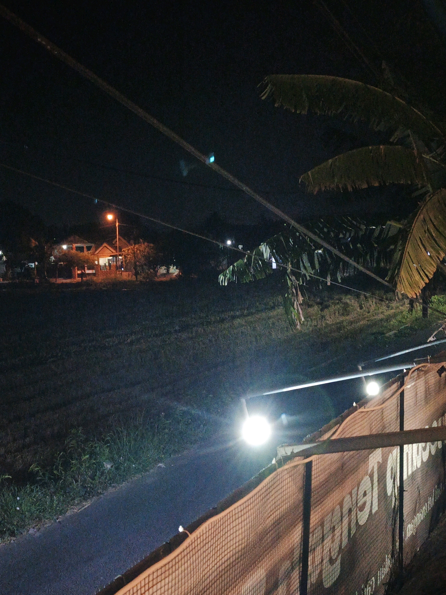 A nighttime, eye-level, medium shot captures a dark, rural scene on a road in Ketegan, Indonesia. The image is illuminated by streetlights and other artificial lights, creating a stark contrast between light and shadow. In the background, a cluster of buildings with lit windows suggests a small residential area or village. Palm fronds from a nearby tree hang over power lines that crisscross the frame. A fence, possibly a temporary barrier, runs along the right side of the road, partially obscuring a large banner with text that is difficult to decipher due to the lighting and angle. The road itself is a dark asphalt surface, bordered by overgrown grass and vegetation. The overall atmosphere is quiet and still, typical of a rural setting at night.