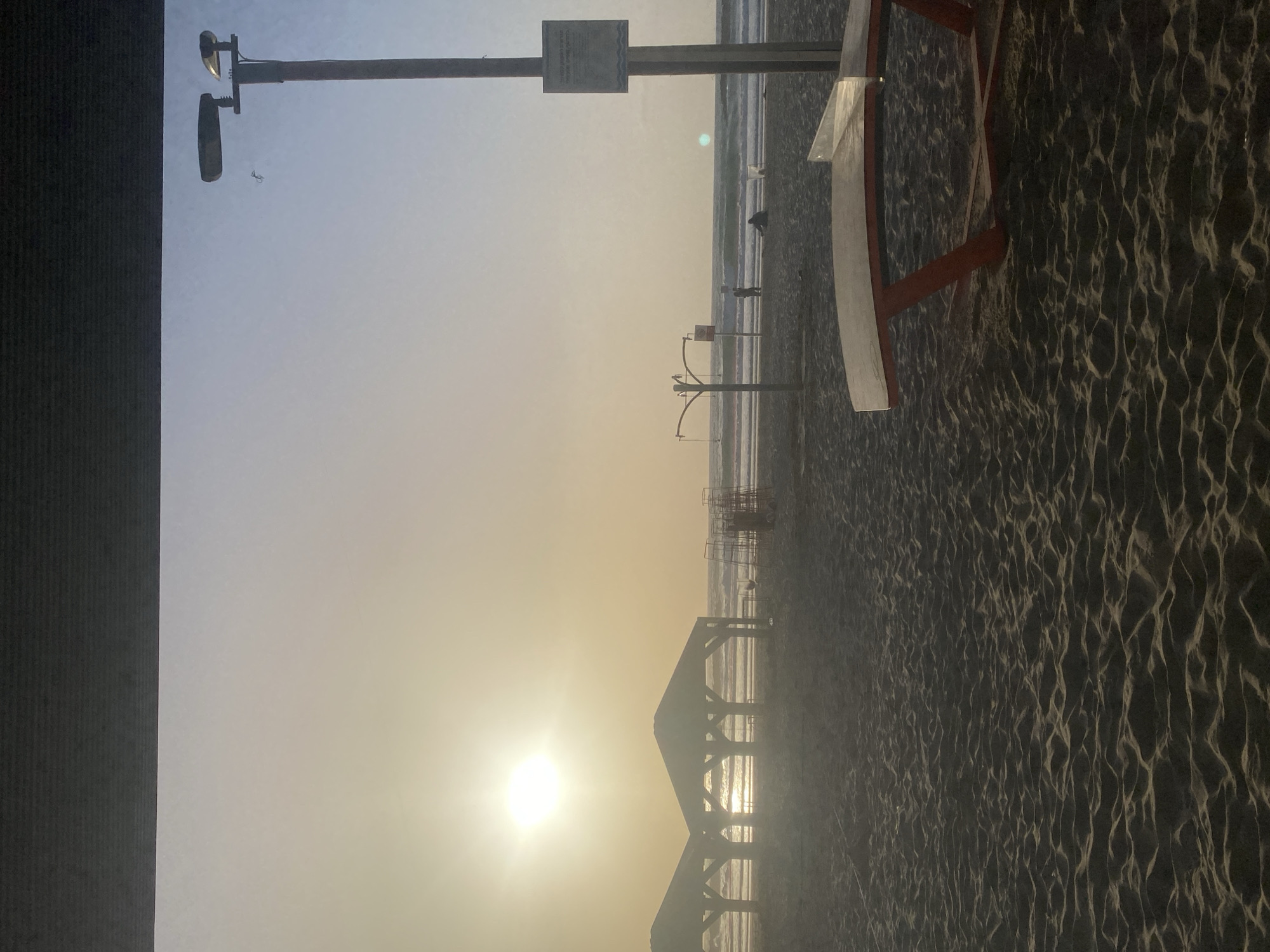 This is a photograph of a beach in Tel Aviv, Israel, likely taken at sunset. The sun is low on the horizon, creating a bright glare. The beach sand stretches across the bottom of the image, showing patterns created by wind or footprints. In the middle ground, there are several structures, possibly lifeguard stands, and silhouetted figures of people walking along the water's edge. In the foreground, there's a partial view of a lifeguard tower and possibly another structure. The sky is bright, transitioning from blue to yellow-orange near the horizon. A light post with cameras is also in the photo.