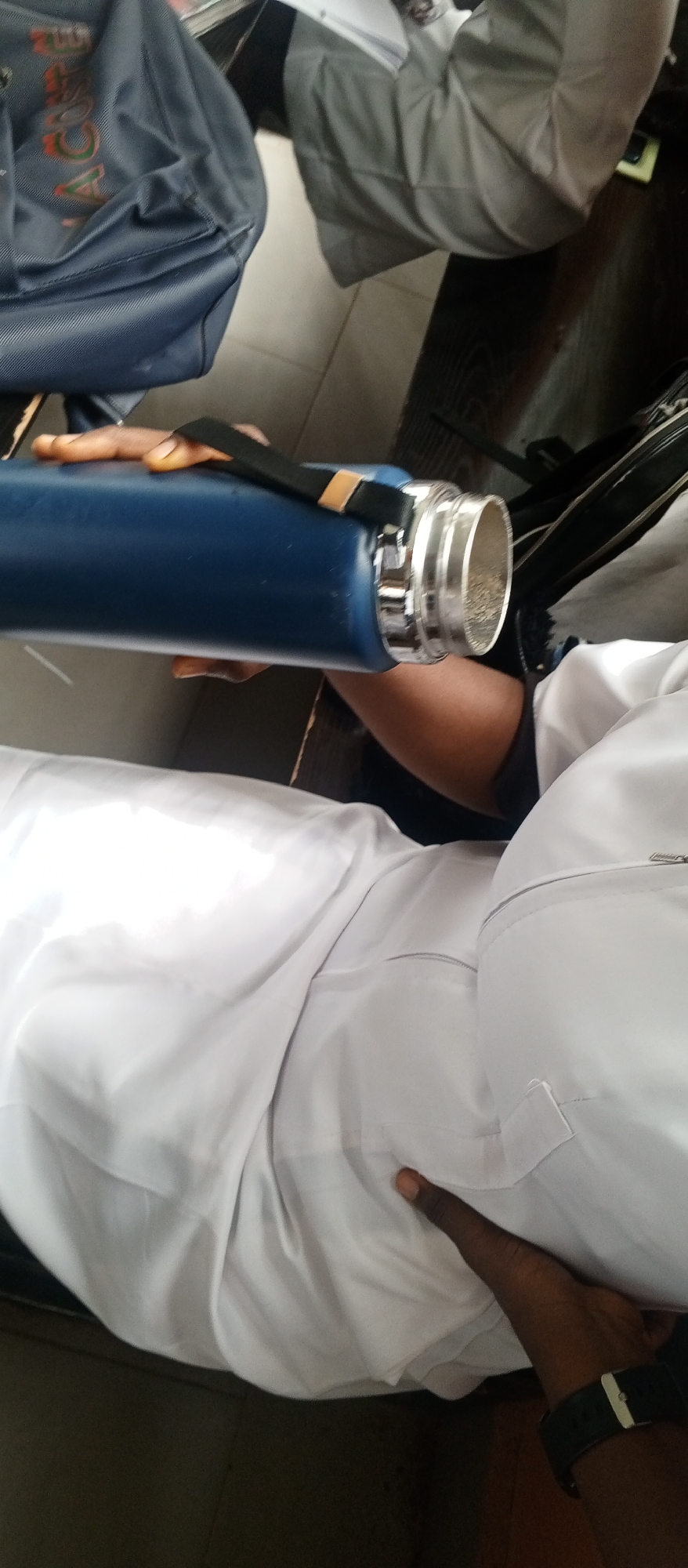 The image shows a person holding a blue water bottle. The person is wearing a white garment, possibly a uniform. In the background, a dark blue bag with some text is visible. The setting appears to be indoors, with tiled flooring. There is no specific information about the location in the city of Kuson, Nigeria, visible in the image.