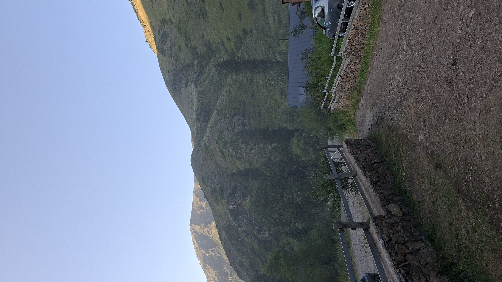 The image depicts a mountainous landscape at dawn, likely in the foothills surrounding Almaty, Kazakhstan. In the foreground, a dirt road curves to the right, flanked by stone walls topped with wooden fences. A dark gray sedan is parked on the right side of the road, near a building with a dark, sloped roof. The middle ground is filled with dense green trees, and the background is dominated by rolling, grassy hills that recede into the distance. The sky is a clear, pale blue, with the sun low on the horizon, casting a warm glow on the upper slopes of the mountains. There are no people visible in the image. The overall impression is one of a serene, natural setting, possibly a rural or semi-rural area near Almaty, known for its proximity to the Tian Shan mountains.