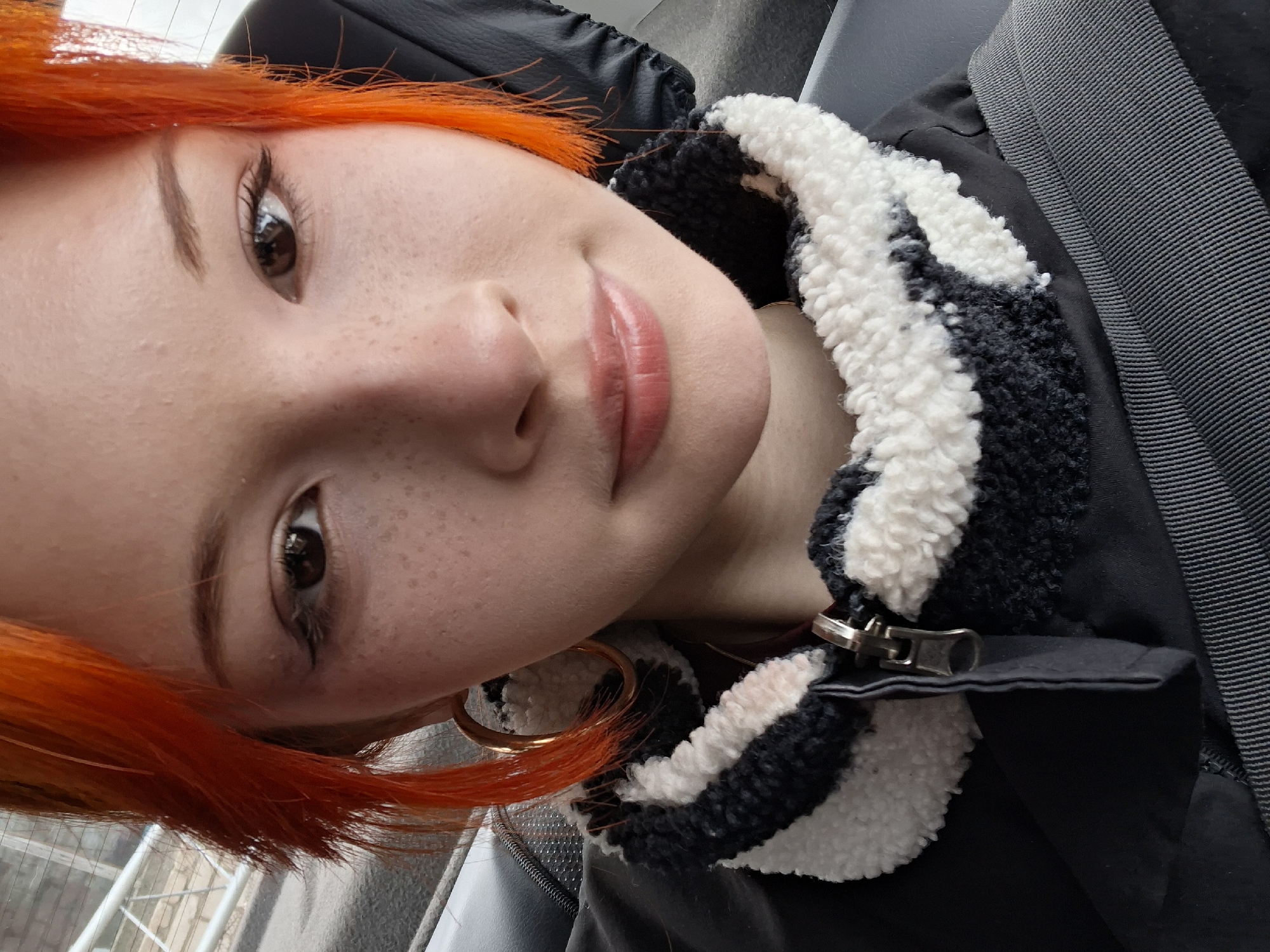 A close-up, selfie-style image features a fair-skinned person with orange hair. The individual's face is partially rotated, presenting a slightly angled view. Their expression appears calm and somewhat serene, with a slight hint of a smile. Freckles are visible across their nose and cheek. The person is wearing a black jacket with a black and white, fluffy collar. They also appear to have a large gold earring in their left ear. A seatbelt is visible across their chest. The background is largely out of focus, with just the suggestion of an exterior scene visible through a window. The location is not discernible from the visual information in the provided image; thus, the city of Chaikovsky, Russia, cannot be confirmed.
