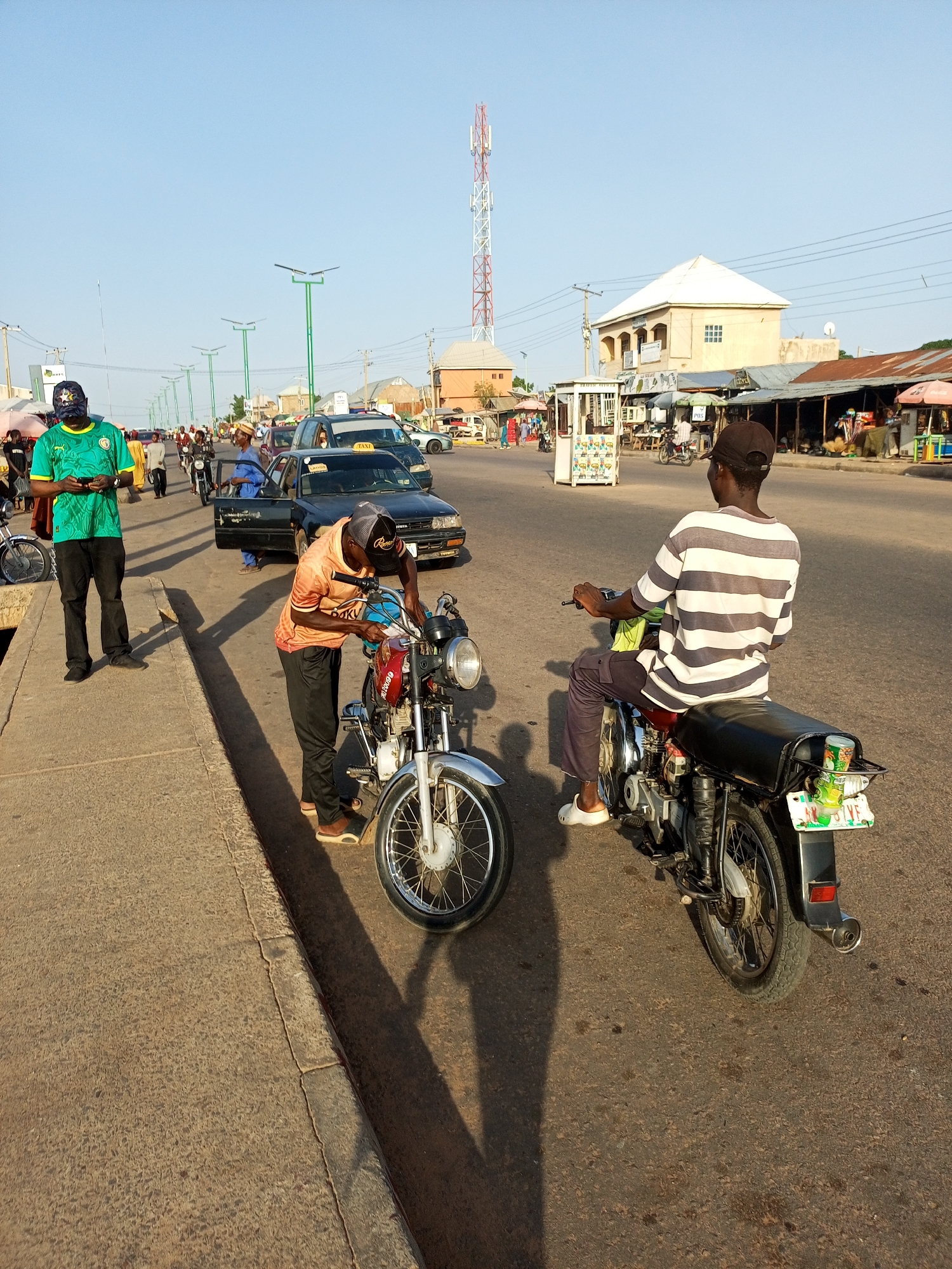 The image depicts a street scene in Tumfure, Nigeria, under a clear sky. In the foreground, two men are interacting with motorcycles. One man, wearing a green shirt and a cap, stands on the left, holding a mobile phone. Beside him is a motorcycle. To the right, another man, dressed in an orange shirt and a cap, is working on a motorcycle. He is bent over, appearing to be adjusting it. A second motorcycle is positioned behind him, with a man in a striped shirt seated on it, facing away from the camera. In the background, traffic including cars and more motorcycles is visible on the road. Buildings and market stalls line the street, suggesting a bustling urban environment. A prominent cell tower stands tall in the distance, and streetlights are spaced along the roadside.