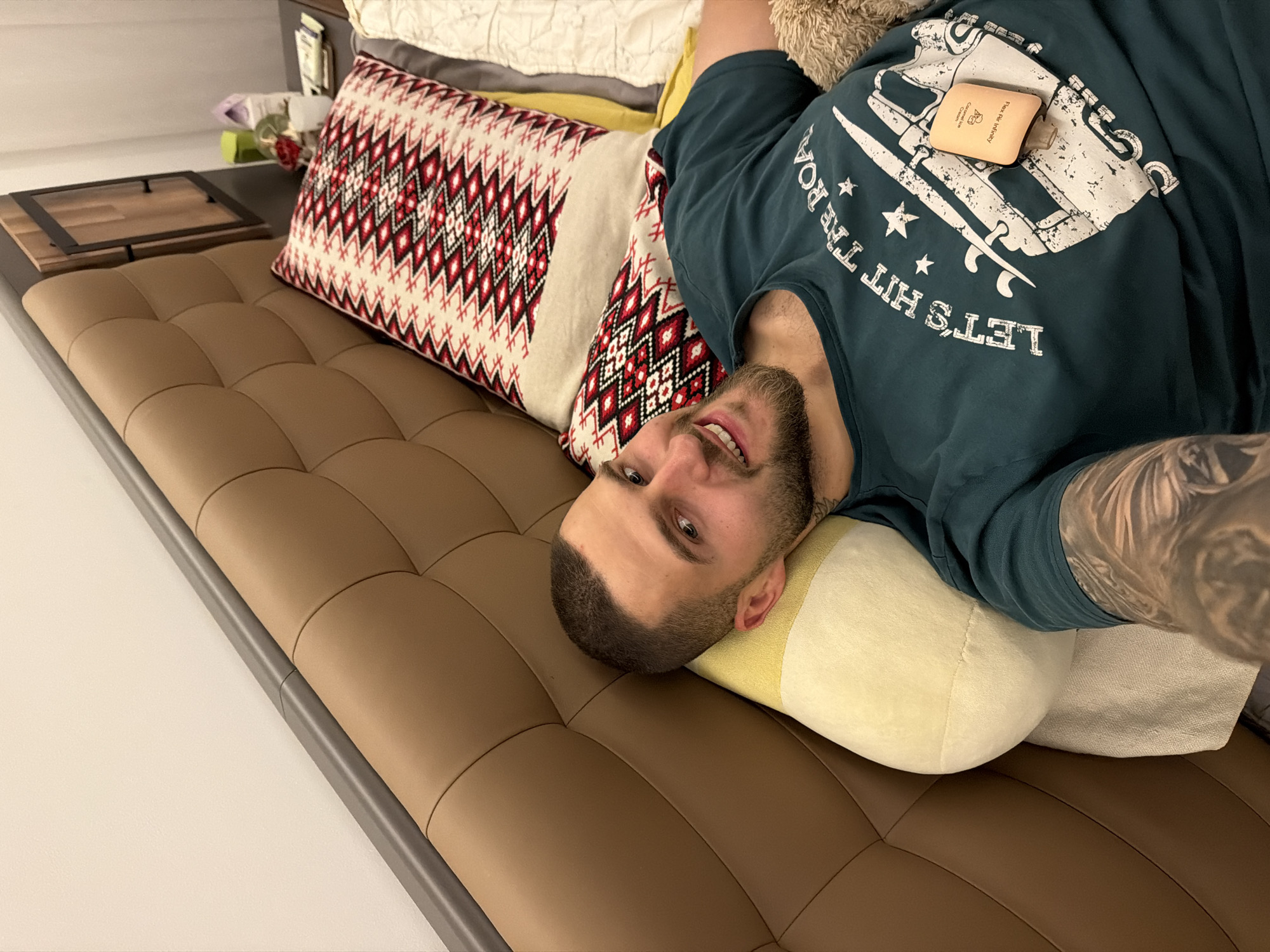 A man with a short buzz cut and a beard is lying on a bed, looking at the camera with a smile. He is wearing a dark green t-shirt with a graphic design and some text. A small orange container is placed on his t-shirt. The bed has a large, tufted brown headboard and is adorned with pillows, one of which has a red, white, and black geometric pattern. To the right of the bed, a nightstand is visible with some items on it, including a red rose in a vase. The overall setting appears to be a bedroom. The image is taken from a high angle, looking down at the man. The location is identified as Sofia, Bulgaria.
