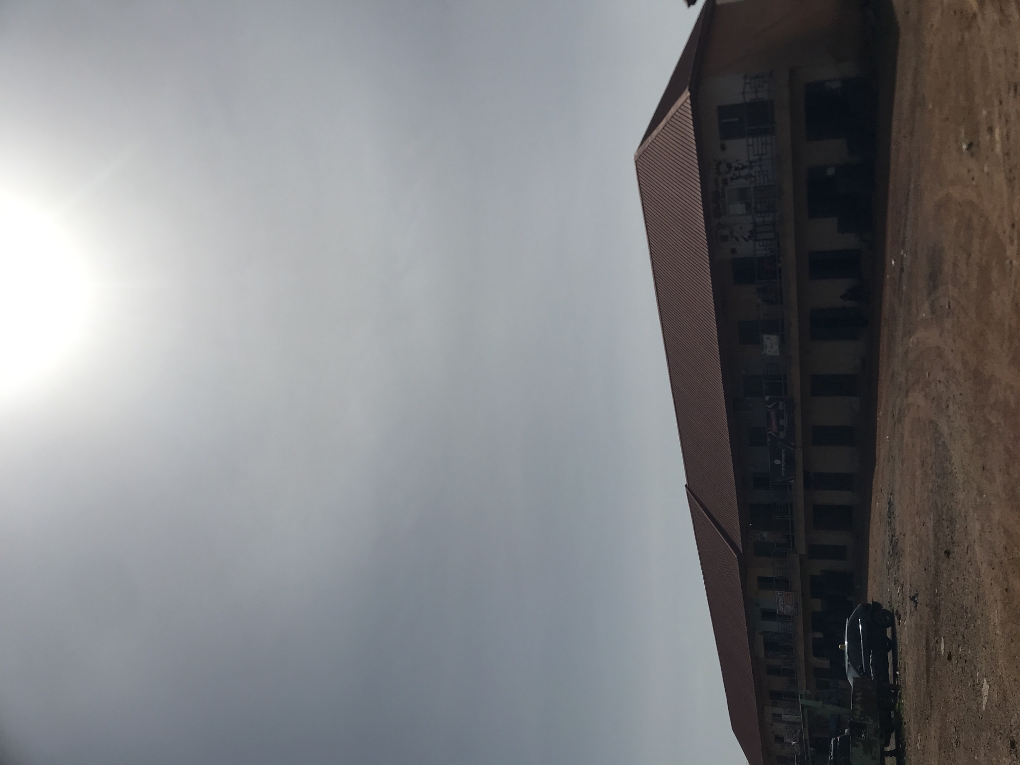 The image captures a daytime scene, originally rotated, dominated by a hazy or overcast sky on the left. A bright, diffused sun is prominently visible in the upper left corner, casting a strong glare. To the right, a long, two-story building stretches horizontally across the frame. It features a reddish-brown sloped roof and light-colored walls. The building's numerous windows appear dark and rectangular. Along the upper floor, scaffolding or metal structures are visible, and several banners with indistinct white text on a dark background hang from the facade.
In front of the building, a reddish-brown dirt ground extends, appearing dry and unpaved. Several dark-colored vehicles are parked on this ground, including a car, possibly a sedan or SUV, towards the right, and another utility-type vehicle partially visible further left. No people are visible in the frame. The bright sun suggests it is daytime, likely late morning or early afternoon, under hazy weather conditions. The setting appears to be a functional complex in a rural or semi-urban environment, fitting the location context of Rafin Par, Nigeria.