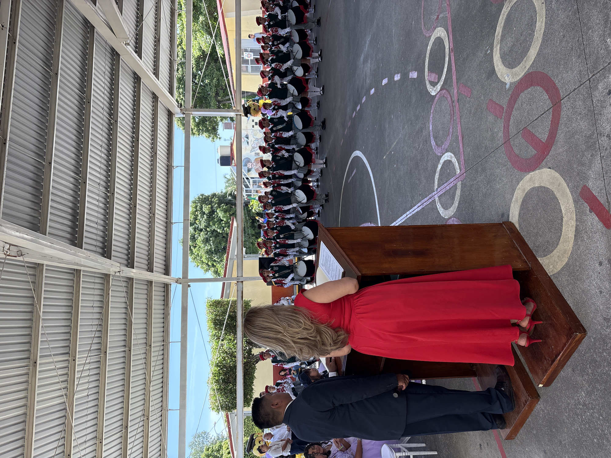 In an outdoor covered area, likely a school courtyard in Tejalpa, Mexico, a formal ceremony is underway. The foreground features a wooden podium with a woman in a vibrant red dress speaking into a microphone. Beside her stands a man in a dark suit. Behind them, a marching band composed of young people in uniform, many wearing red berets and dark vests over white shirts, stands at attention, holding drums. The ground is a paved surface with painted markings. In the background, buildings and lush green trees are visible under a bright sky. The overall scene suggests an event involving a school band and a public address.