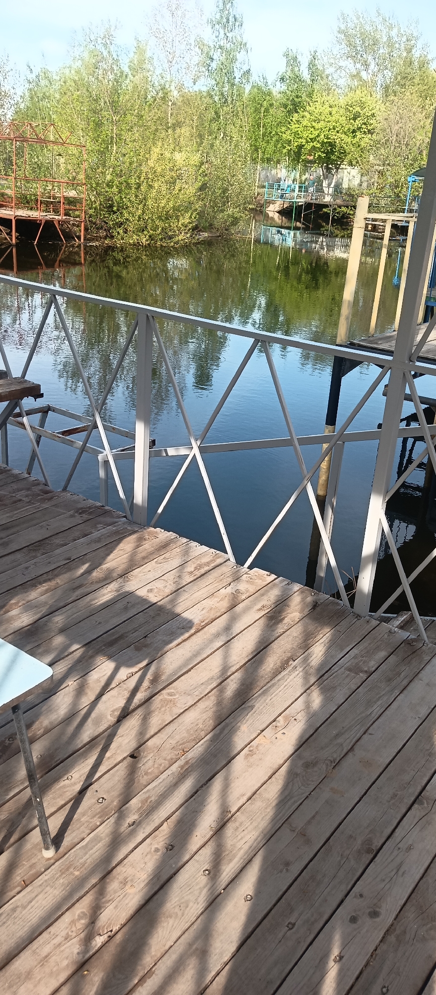 The image shows a wooden deck with a white metal railing overlooking a body of water. Beyond the water, there are trees and structures, possibly other docks or buildings. A table is partially visible on the deck. The image has an outdoor setting under daylight conditions. The specific location is not discernible from the image.