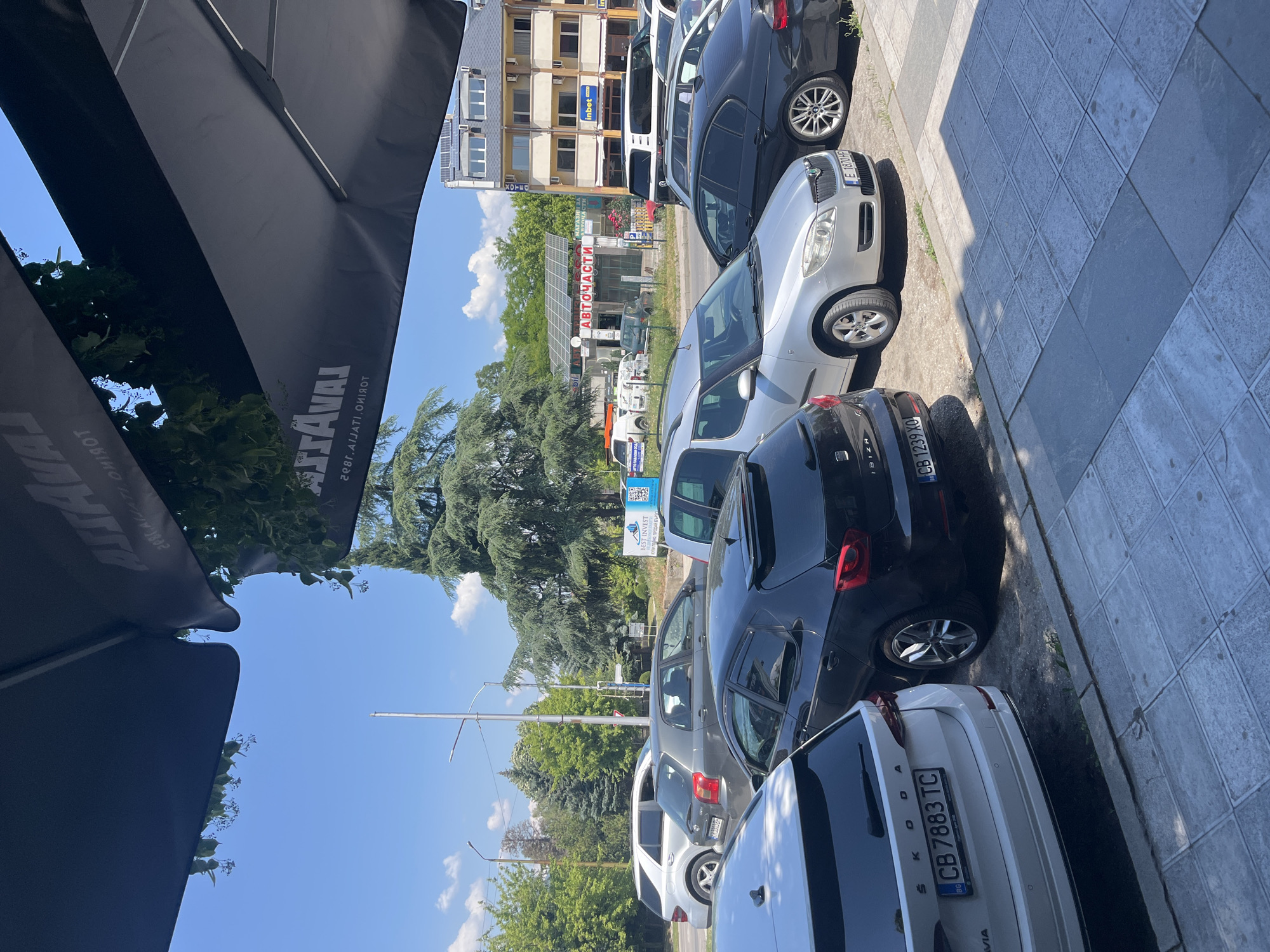 Here's a matter-of-fact description of the image:
The image depicts a street scene with multiple parked cars along a sidewalk. The cars are of various makes and models. License plates are visible on some of the cars.
Behind the parked cars, there's a row of trees and beyond that, a multi-story building. Signs written in a non-English language are visible on the building.
An umbrella is partly visible in the foreground, obscuring part of the sky.