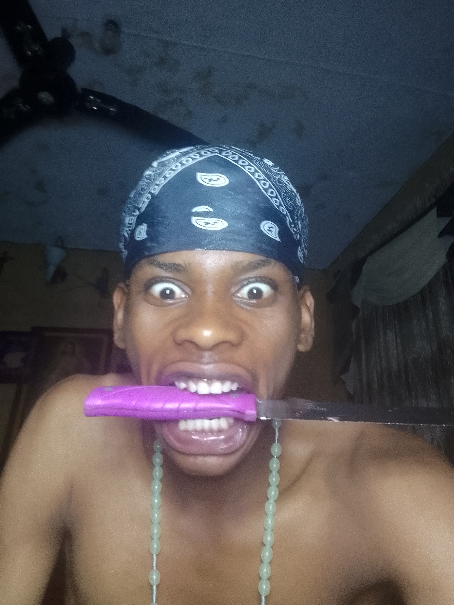 A shirtless person with dark skin and a blue paisley bandana holds a knife in their mouth. The knife has a pink handle and a silver blade. The person's eyes are wide. They wear a string of light green beads around their neck. A dark ceiling fan is visible in the upper left of the frame. A blurred picture frame hangs on a wall in the background. The location is Unknown, Unknown.