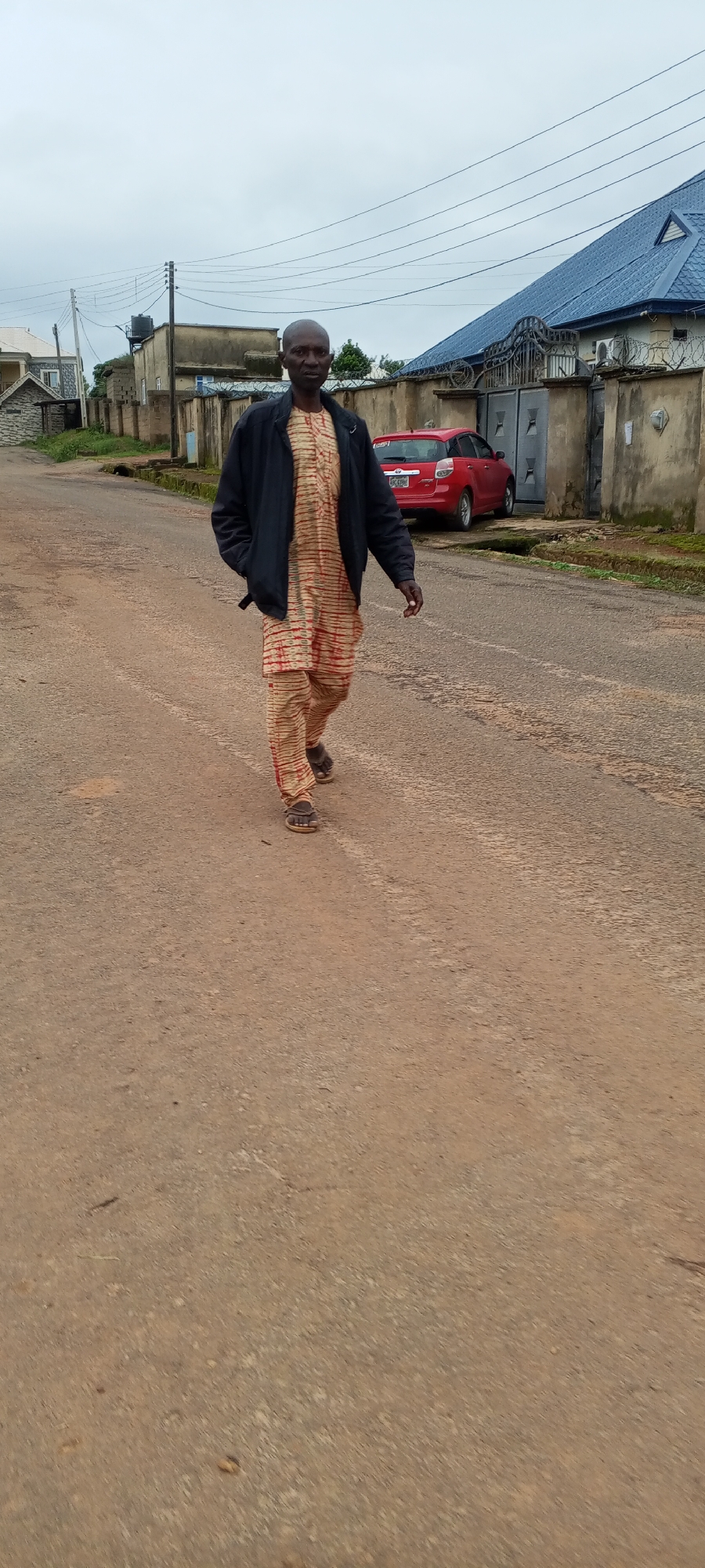An adult male walks on a dirt road in front of a house with a blue roof and a large metal gate. He wears a dark jacket over a patterned tunic and trousers and brown sandals. A red car is parked on the road behind him. The sky is overcast. The scene is likely in Bahit, Nigeria.