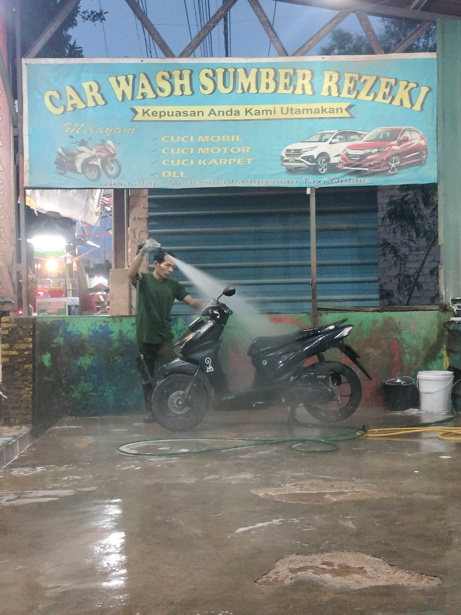The image depicts an outdoor car and motorcycle wash establishment in Batam, Indonesia. A man in a green t-shirt is actively washing a black motorcycle with a high-pressure spray. The backdrop features a blue banner with yellow lettering that reads "CAR WASH SUMBER REZEKI," indicating the name of the business. Below the name, the banner lists services offered, including "CUCI MOBIL" (car wash), "CUCI MOTOR" (motorcycle wash), "CUCI KARPET" (carpet wash), and "DLL" (and so on). It also states "Kepuasan Anda Kami Utamakan" (Your Satisfaction is Our Priority) and "Bisa Juga Menerima Langganan Taxi Online" (Also Accepts Online Taxi Subscriptions). In the background, there are images of a white car and a red car, suggesting the types of vehicles serviced. The ground is wet and appears to be concrete, with puddles of water and some soapy residue. Two buckets, one black and one white, are visible near the washing area, along with a green and a yellow hose. The overall environment appears to be a functional, somewhat utilitarian space for vehicle cleaning.