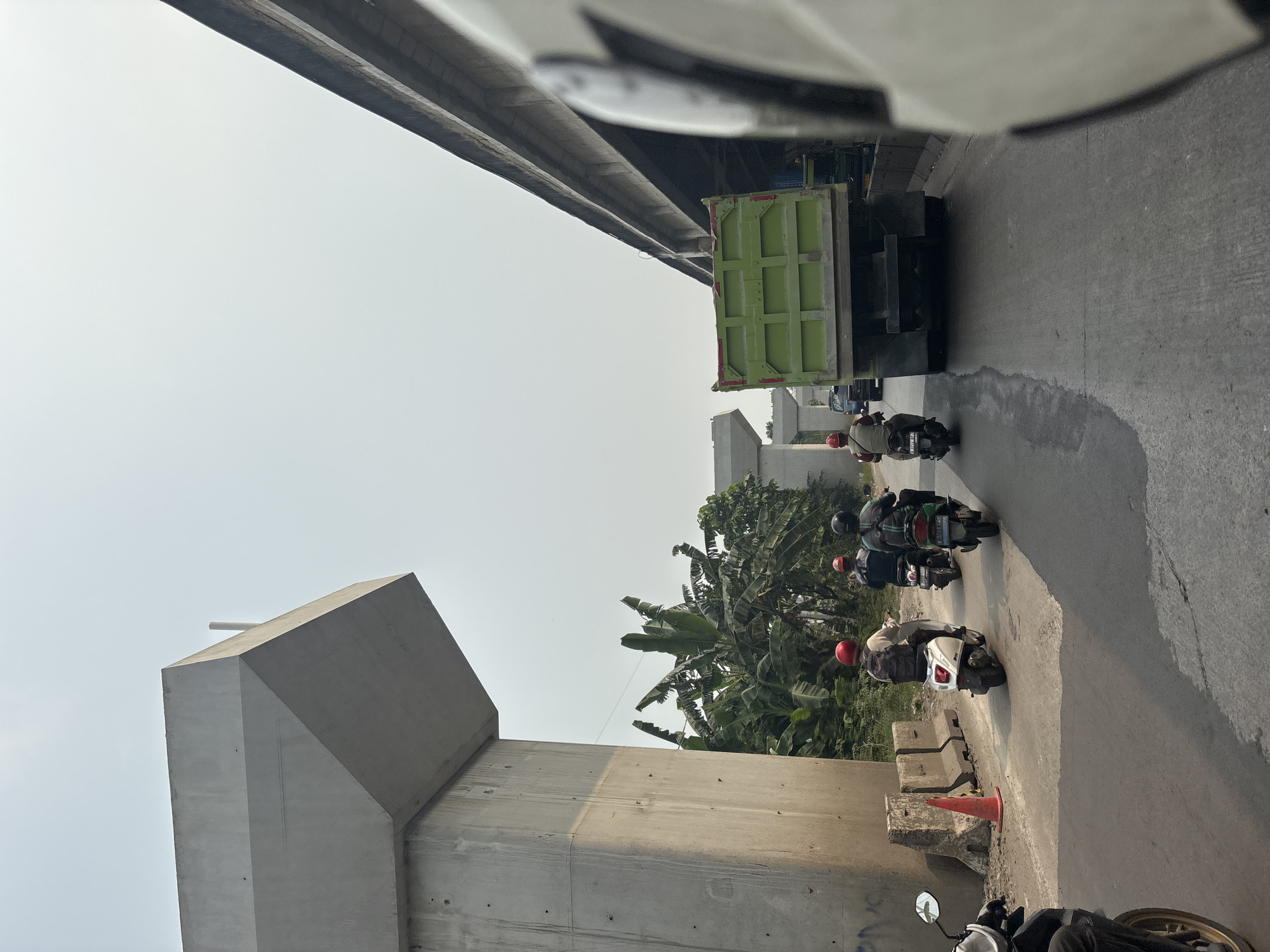 In North Jakarta, Indonesia, a daytime view captures traffic on a road under construction, evidenced by large concrete structures for an elevated highway or bridge. The scene is dominated by the imposing concrete pillars and beams of this infrastructure project. To the left, a massive, angular concrete pillar stands tall, casting a shadow on the road. Further back, similar concrete supports are visible, suggesting ongoing construction.
The road itself is paved and appears to be a busy thoroughfare. Several motorbikes are present, navigating the traffic. A person on a white scooter, wearing a red helmet and a backpack, is moving away from the camera. Behind them, two more individuals on motorbikes, also wearing helmets, are traveling in the same direction. In the distance, a large green dump truck is a prominent feature, positioned in the middle of the road. A dark-colored vehicle is visible behind the dump truck.
To the left of the road, a cluster of banana trees and other lush green vegetation creates a natural contrast to the man-made concrete structures. A bright orange traffic cone sits on the side of the road near some concrete barriers. The sky above is bright and clear, indicating fair weather. The overall impression is of a developing urban environment, where modern infrastructure construction intersects with daily life and traffic.