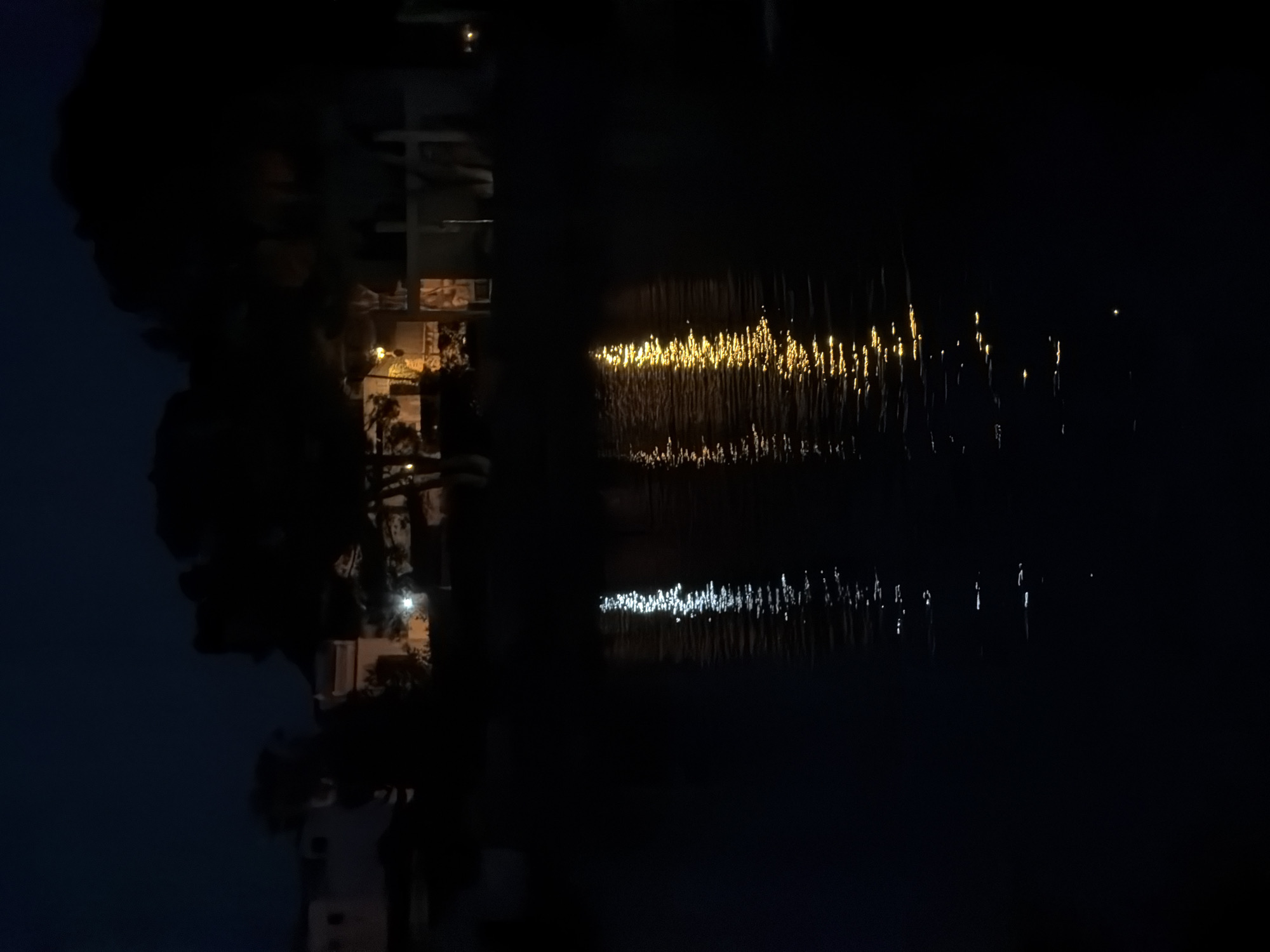 The image depicts a nighttime scene in Marina Manresa, Spain, showing a body of water reflecting the lights of buildings and streetlights. The reflections create a streaked, glittering effect on the water's surface. The buildings are silhouetted against the dark sky, with illuminated windows visible. Trees are present on the shoreline, their shapes appearing as dark masses. The overall scene evokes a sense of tranquility and urban night ambiance.