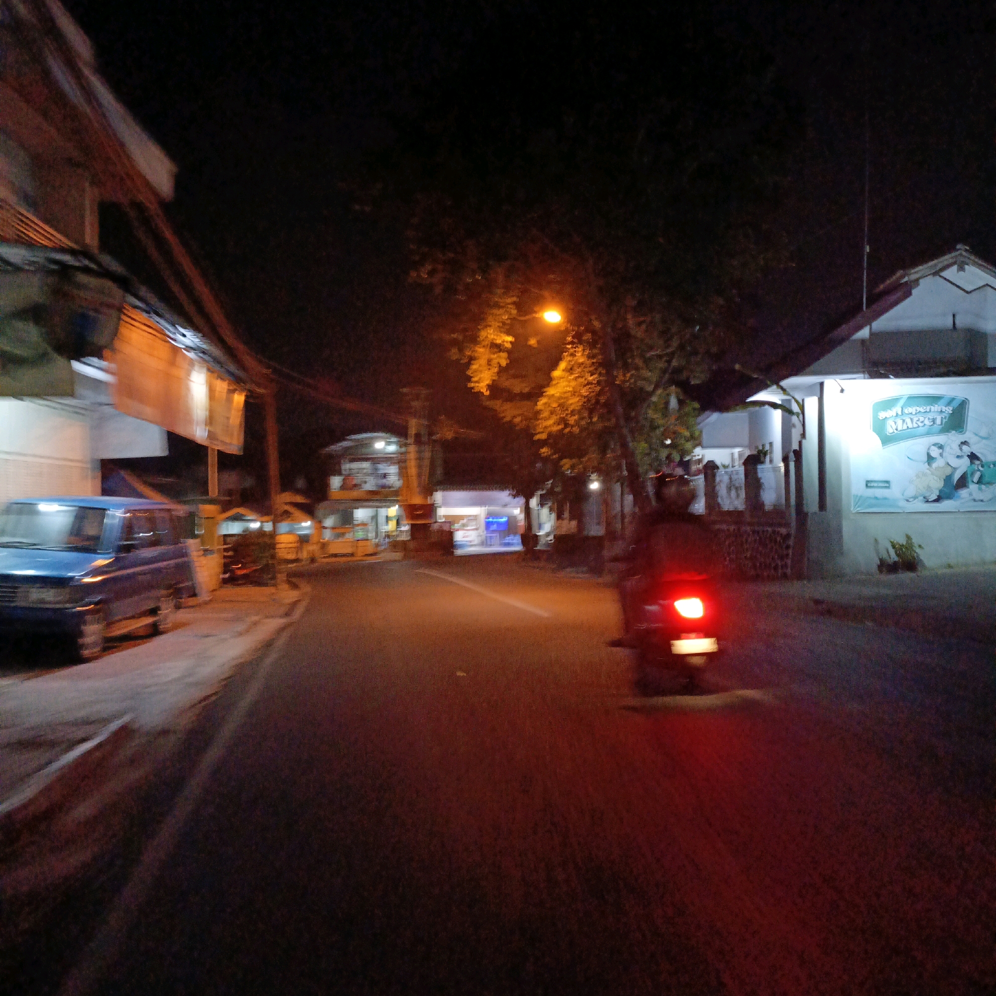 In the city of Salatiga, Indonesia, at night, a street scene unfolds with a blue van parked on the left side of the road. A street lamp with an orange glow illuminates the trees and buildings. On the right side of the road, a motorcycle is in motion, its red taillight reflecting on the asphalt. Buildings line both sides of the street, some with lit windows and signage. One building on the right features a large green and white banner with text that appears to say "soft opening MARET." The overall atmosphere is dimly lit, characteristic of a nighttime street scene in a residential or commercial area.
