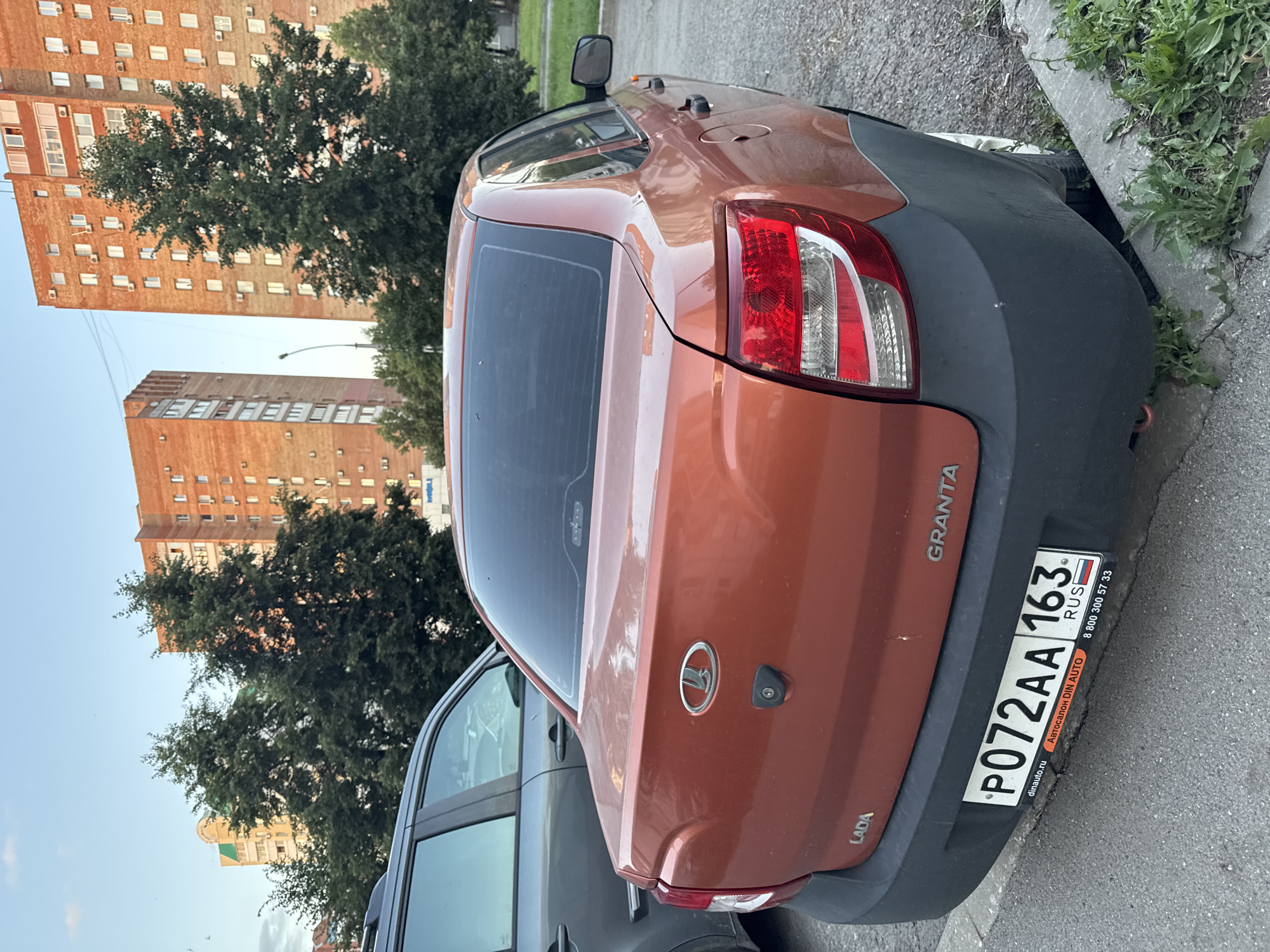 An orange Lada Granta sedan is parked on a gravelly shoulder next to a paved road. The car is facing away from the camera and is slightly angled to the right. A dark grey SUV is parked to the left of the Lada. In the background, two tall, multi-story brick apartment buildings stand under a clear sky with a few wispy clouds. Green trees are visible in front of the apartment buildings and to the right of the car. The license plate of the Lada reads "P072AA 163". The car appears to be parked in a residential area.