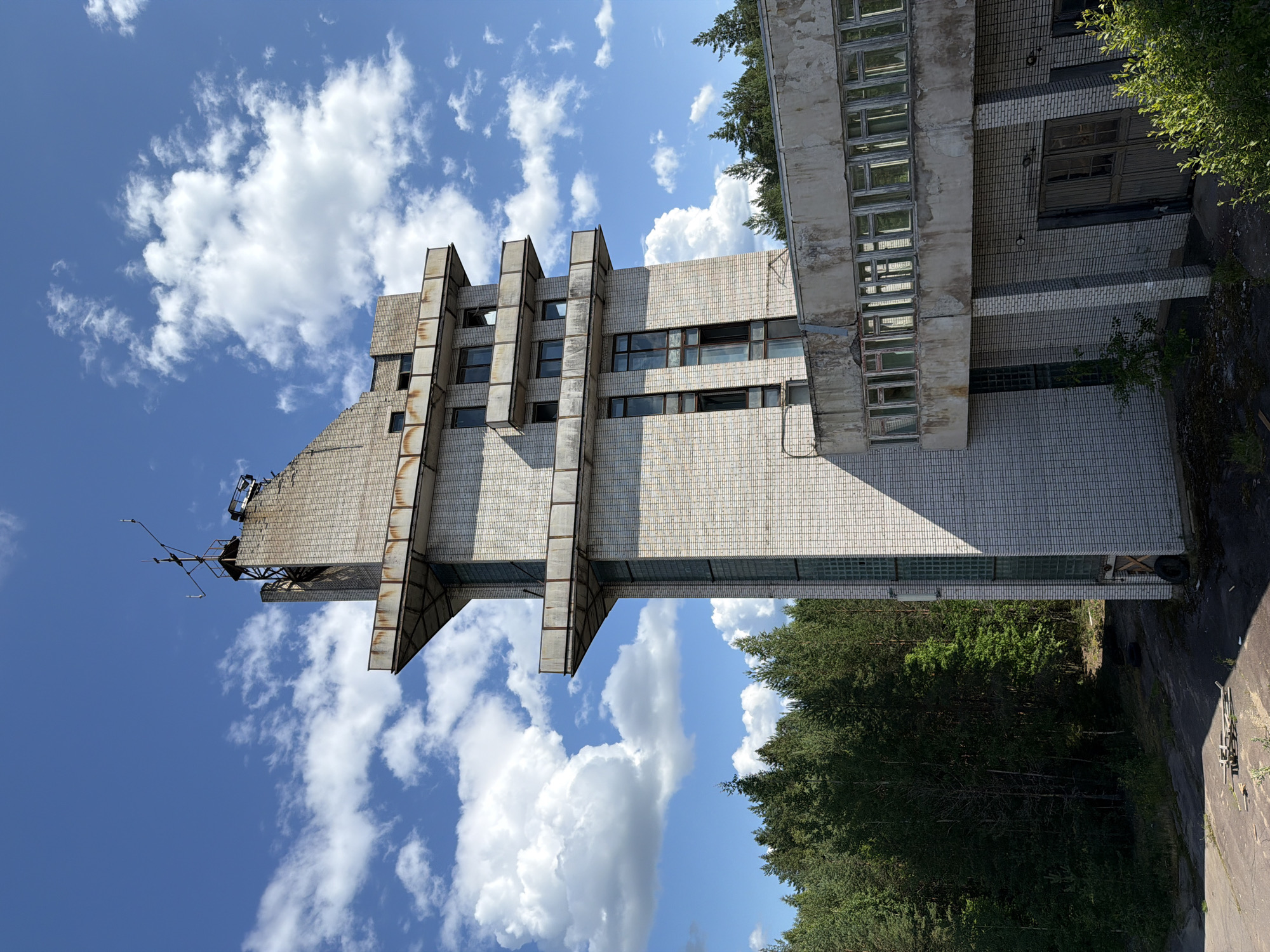 This image shows an abandoned, multi-story building in Nikolsky, Russia, set against a backdrop of a blue sky with white clouds and a dense forest. The building is constructed of light-colored bricks and features several balconies with metal railings. The architecture is angular, with some sections appearing to jut out. The windows are numerous, and many of them appear broken or boarded up. At the top of the tallest section of the building, there is a structure resembling an antenna or communication tower. The ground around the building is covered in patchy vegetation and debris, suggesting neglect. The overall scene conveys a sense of abandonment and decay.