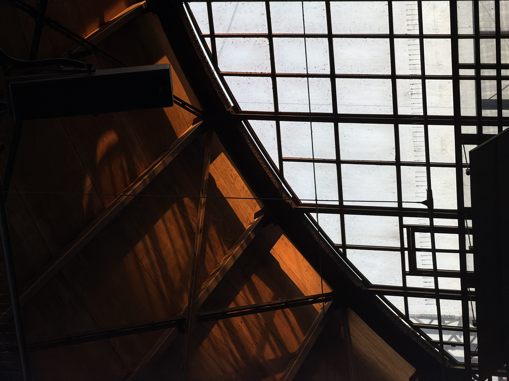 The image captures the interior of a large, industrial-style building, possibly a former factory or industrial hall, located in Krefeld, Germany. The architectural style suggests a historical industrial site, possibly an early 20th-century structure given the combination of wood and metal construction.
The dominant feature is the large, arched window system that fills the right side of the frame. These windows are divided into numerous panes by dark metal mullions, and the glass itself is textured or frosted, diffusing the light that enters. The windows are set into a substantial steel framework.
Above the windows, the underside of a wooden roof structure is visible. This structure is supported by large metal beams and trusses, creating a complex geometric pattern. Sunlight streams through gaps or the upper portion of the roof, casting strong, sharp shadows from the beams onto the wooden ceiling. The light hitting the wood has a warm, orange hue, indicating it is likely late afternoon or early morning sunlight.
A black, rectangular speaker hangs from the ceiling near the top center of the image, suspended by wires. A thin, dark string or wire also hangs down from the ceiling in the middle ground, extending towards the window area.
In the lower part of the image, dark silhouettes of interior structures are visible, including what appears to be a railing or walkway and possibly the edge of a mezzanine or upper floor. These elements are obscured by shadow, adding to the sense of depth and scale within the building. The overall impression is one of an atmospheric and somewhat stark industrial space, illuminated by dramatic natural light.