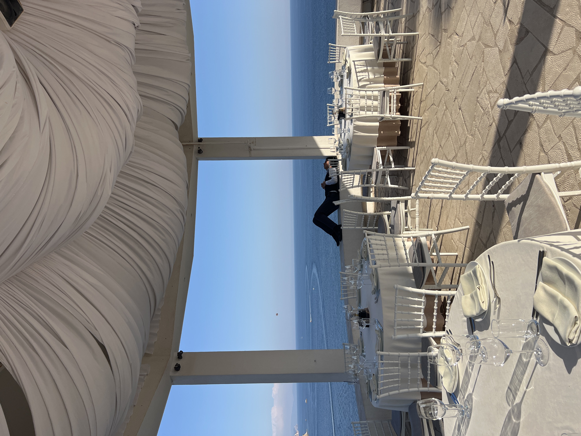 A man in a dark suit and white shirt is lying on his back with his eyes closed on a low wall overlooking the Adriatic Sea. He is situated on the rooftop terrace of a restaurant in Budva, Montenegro, evidenced by the distinctive white draped canopy overhead and the blue waters of the sea. The terrace is set for a formal event, with multiple round tables covered in white tablecloths and adorned with place settings, including white plates, napkins, and glassware. White chairs with padded seats surround the tables. The background features a clear blue sky and the calm sea, with a few boats visible in the distance and a parasailer in the air. The flooring of the terrace is made of irregularly shaped stone tiles.