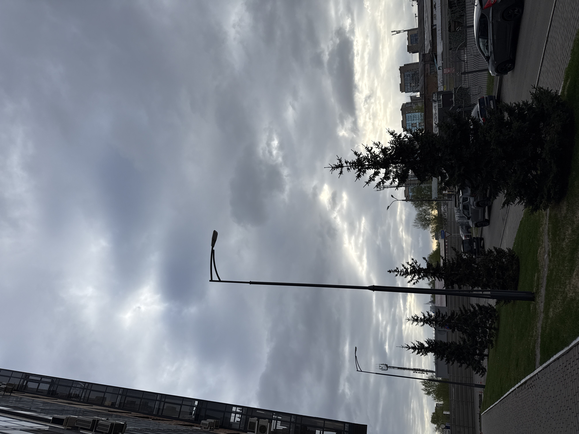 The image shows an urban scene under a cloudy sky. The location is likely Krasnoyarsk, Russia, due to visual cues such as Cyrillic writing and architectural styles typical of the region. The foreground features a modern building with mirrored glass windows. Streetlights are visible extending upwards into the sky. Evergreen trees stand in a grassy area alongside a sidewalk. A street with parked cars is positioned to the right of the trees. Further in the background, buildings are visible, completing the urban landscape. The sky is overcast with thick cloud cover obscuring direct sunlight.