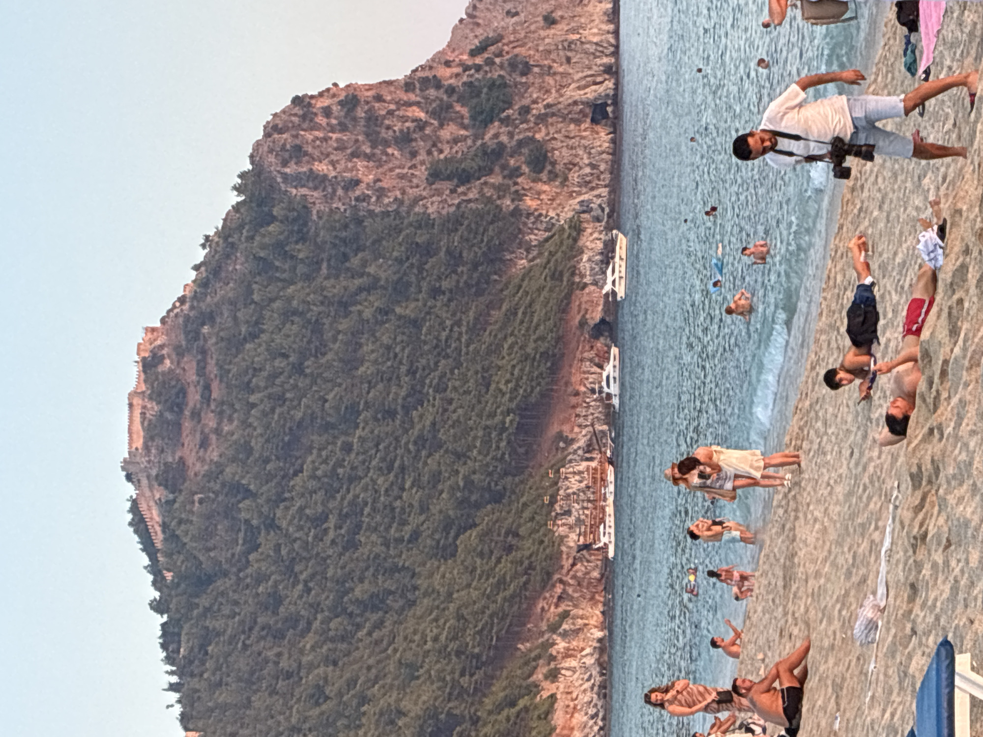 The image captures a coastal scene in Alanya, Turkey, dominated by a dramatic cliff face topped with an ancient fortress. The sea is a calm blue, with several boats, including yachts and a more traditional vessel, anchored offshore. People are scattered across a sandy beach, enjoying the water and the sun. Some are swimming or wading in the sea, while others are relaxing on the sand, sunbathing, or engaged in conversation. In the foreground, a man is standing with a professional camera around his neck, seemingly taking photographs. The overall impression is of a popular tourist destination, showcasing both natural beauty and historical landmarks.