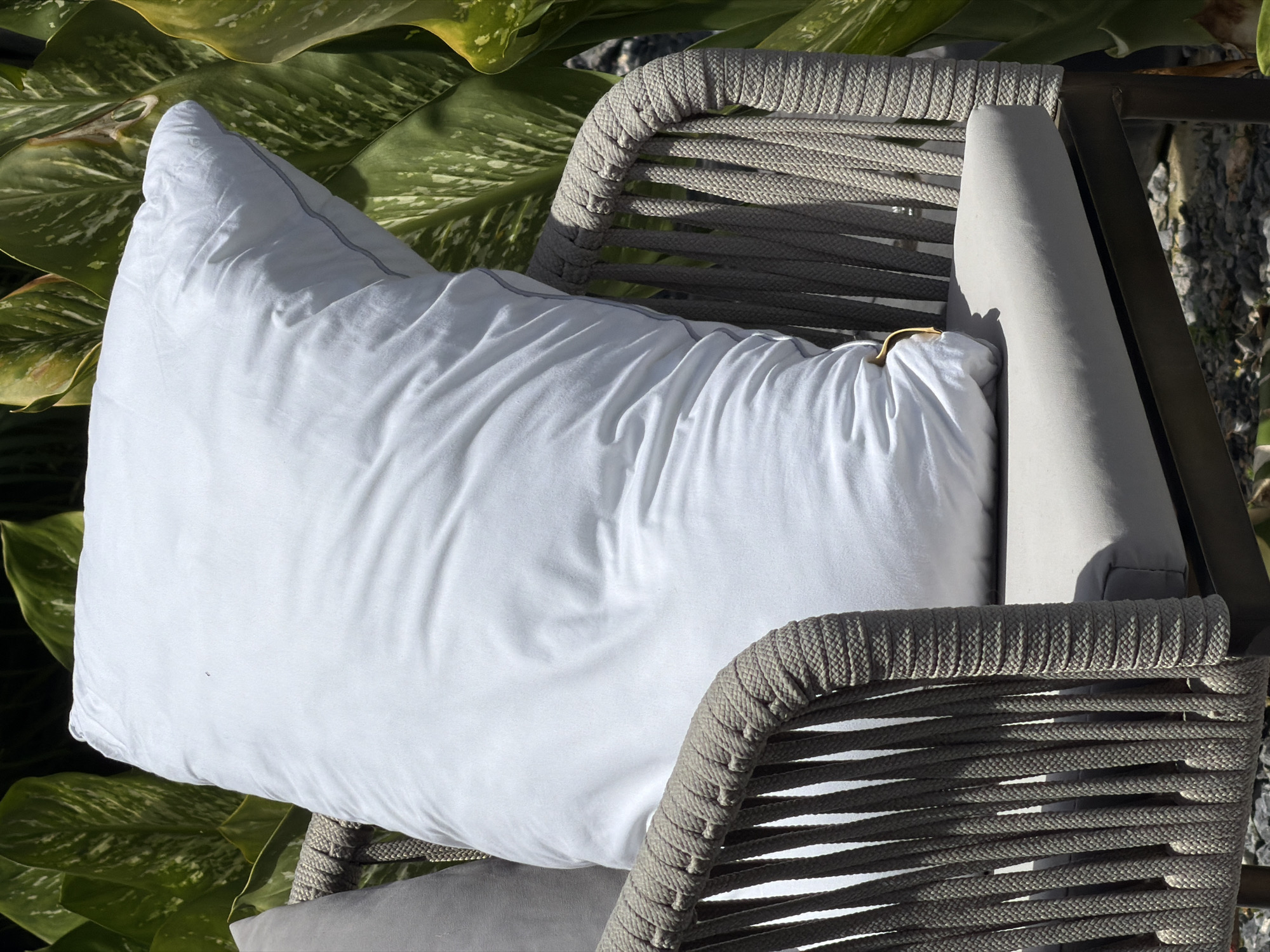 A white pillow is placed on a gray cushioned chair. The chair has a woven rope design on its sides and back. The chair is situated outdoors with lush green foliage in the background. The ground beneath the chair appears to be covered in gravel.