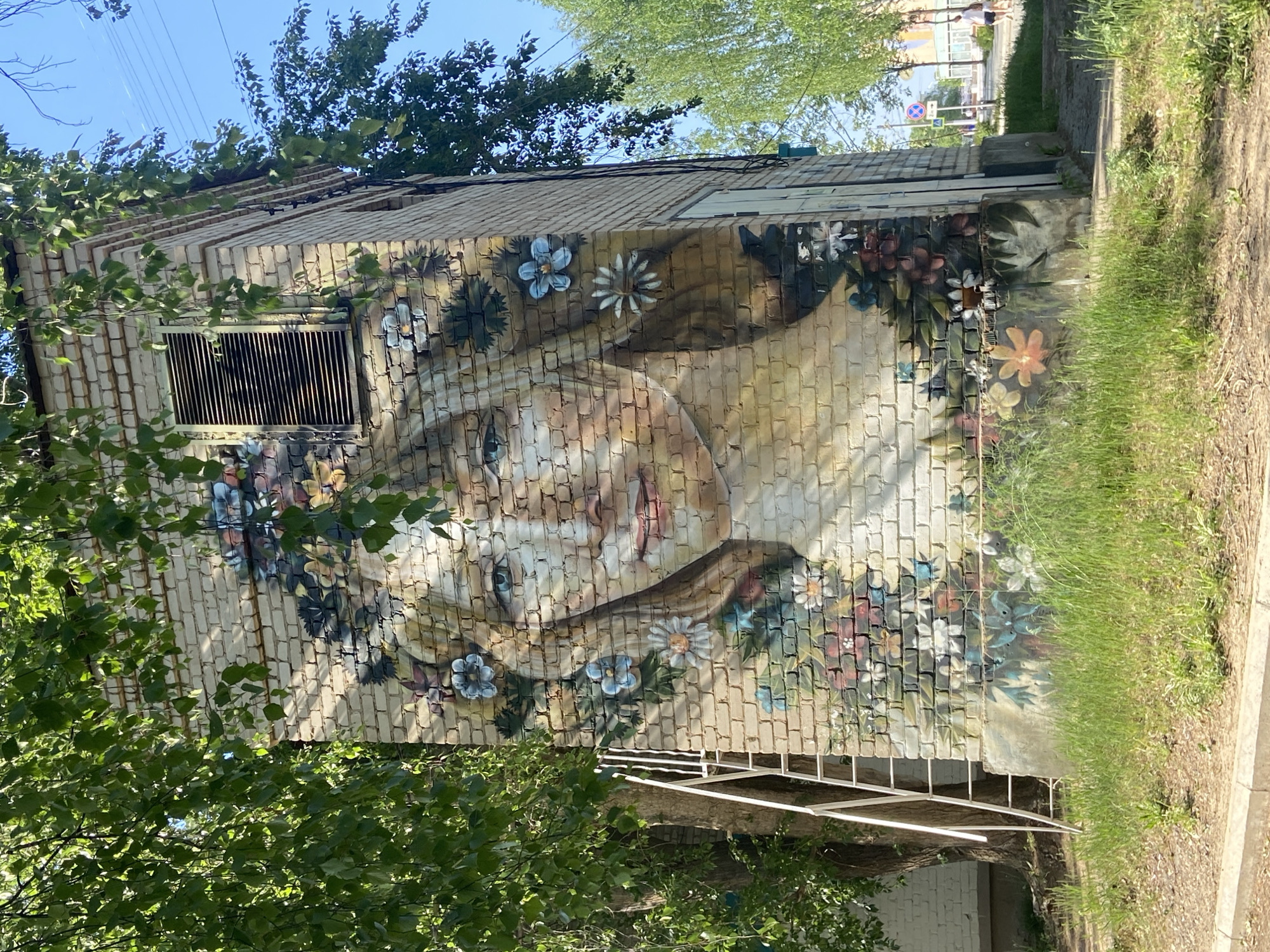 A mural depicting a woman's face surrounded by flowers is painted on the side of a brick building. The building also has a barred window. Grass grows along the building's base, and a sidewalk is visible. Trees frame the building. The location is Chaikovsky, Russia. A section of a metal fence or railing is also visible near the base of the building.