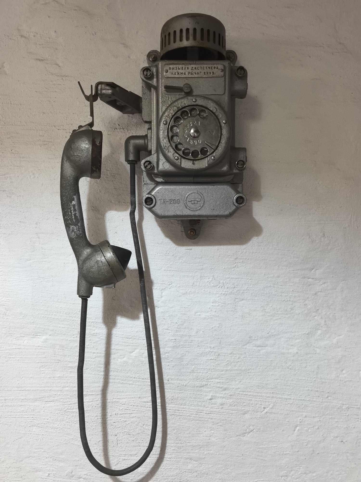 A vintage, wall-mounted industrial or military telephone is the central subject of this image, set against a textured, off-white wall. The telephone, likely made of metal and painted grey, is prominently displayed in the upper-middle section of the frame.
The phone's main body features a circular rotary dial with numbers 0-9 visible, indicating an older communication technology. Above the dial, a grille for a bell or speaker is integrated into the top of the unit. Below the dial, a small rectangular plate bears the model number "ТА-200" and a distinct, stylized logo. The telephone handset hangs freely to the left of the main unit, suspended from a metal hook. It is connected to the base by a dark, coiled cable that forms a U-shape below the handset. The handset itself appears to be of the same metallic material as the base, with black components for the earpiece and mouthpiece.
No activity is taking place; the phone is idle. The setting appears to be indoors, with a light source casting a noticeable shadow of the telephone and its cable onto the wall, indicating depth and texture. The wall itself has a rough, uneven surface, possibly concrete or painted brick.
Visible text on the telephone, written in Cyrillic, translates to instructions: "ВЫЗЫВАЙ ДИСПЕТЧЕРА," (Call the dispatcher,) and "НАЖМИ РЫЧАГ ВНИЗ." (press the lever down.). The model number "ТА-200" is also clearly legible. The phone's design and condition suggest it is a robust, utilitarian device, showing signs of age and wear. This object is located in Chaikovsky, Russia.