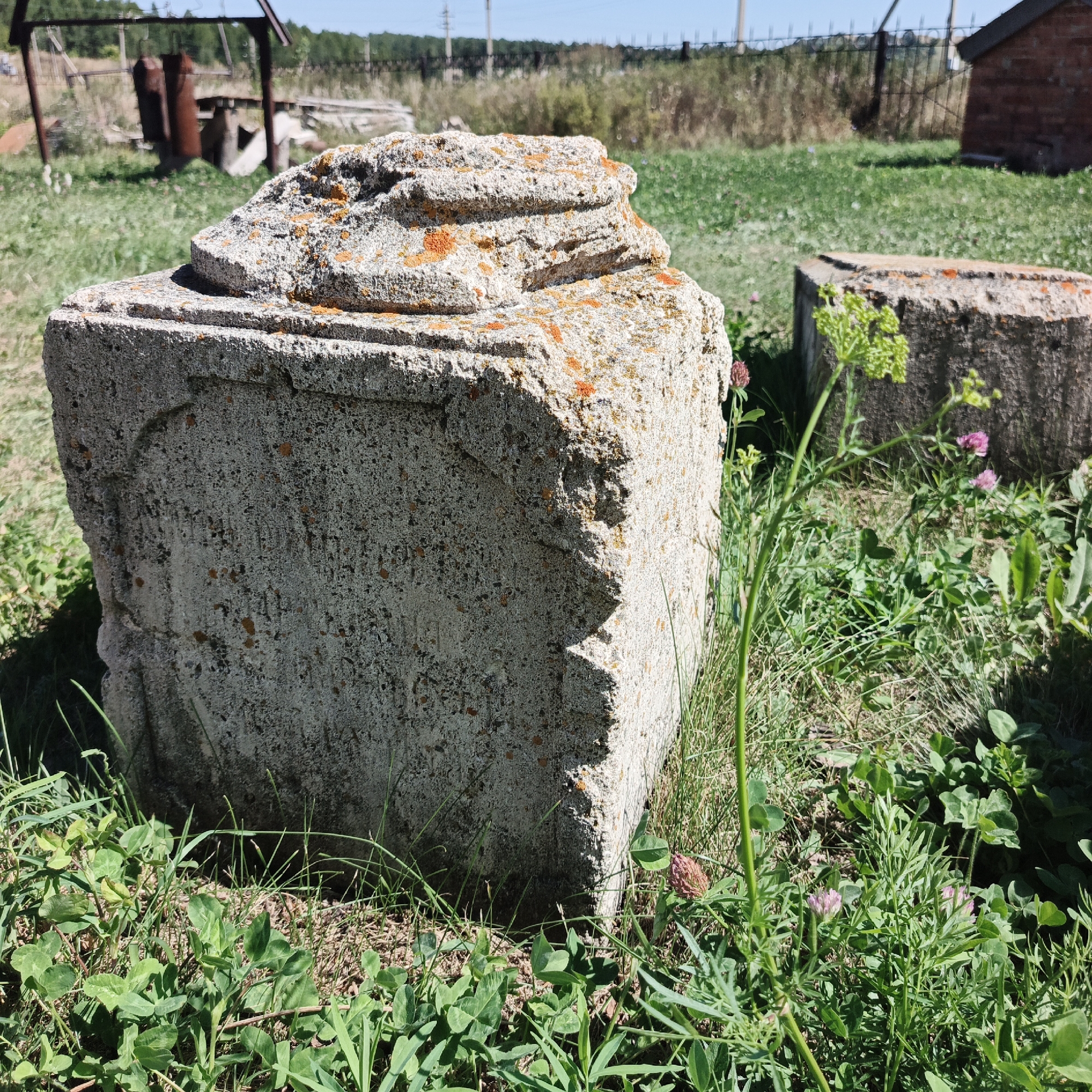 The image depicts two weathered, concrete blocks in a rural, outdoor setting. The foreground is dominated by a large, roughly cubic concrete block with a decorative, layered top, showing signs of age and wear. Orange lichen is visible on its surfaces. To the right, a second, similar concrete block is partially visible, also weathered and with lichen. Both blocks are surrounded by green grass and various small plants, including clover and some with small pink flowers. In the background, a field with dry grass and some scattered vegetation stretches towards a fence and utility poles. A rusty metal structure and some discarded items are visible further in the distance, suggesting an abandoned or undeveloped area. The sky is clear and blue. There are no people or animals present in the image. The scene is consistent with an outdoor location in the village of Tugutustemir, Russia.