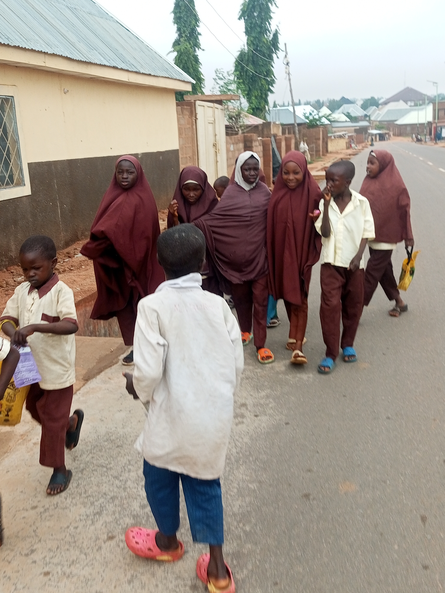 A group of children are walking on a paved road with buildings in the background. A few of the children are wearing maroon hijabs and robes. One child in the foreground is wearing a white long-sleeved shirt and blue pants, and another child is wearing a cream-colored polo shirt with maroon accents and maroon pants. One child is wearing a white shirt and maroon pants, and a pink crocs. Another child is wearing a white shirt and blue pants, and pink crocs. The buildings appear to be houses and a shop. The sky is overcast. The scene is located in Unknown, Unknown.
