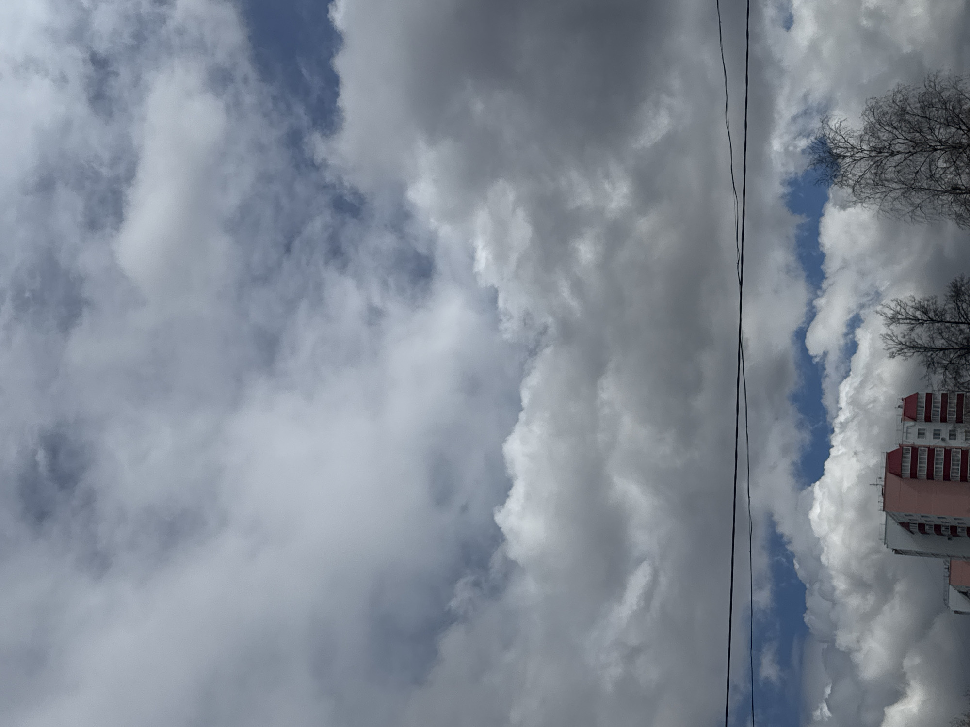 The image shows a view of the sky with heavy clouds, interspersed with patches of blue sky. A power line runs vertically through the scene. In the bottom right corner, a residential building with a red roof is visible. Based on the architectural style, the image is likely taken in a city in Russia. Likely, Perm, Russia, given the prompt. The trees are bare, suggesting it may be late autumn or early spring.