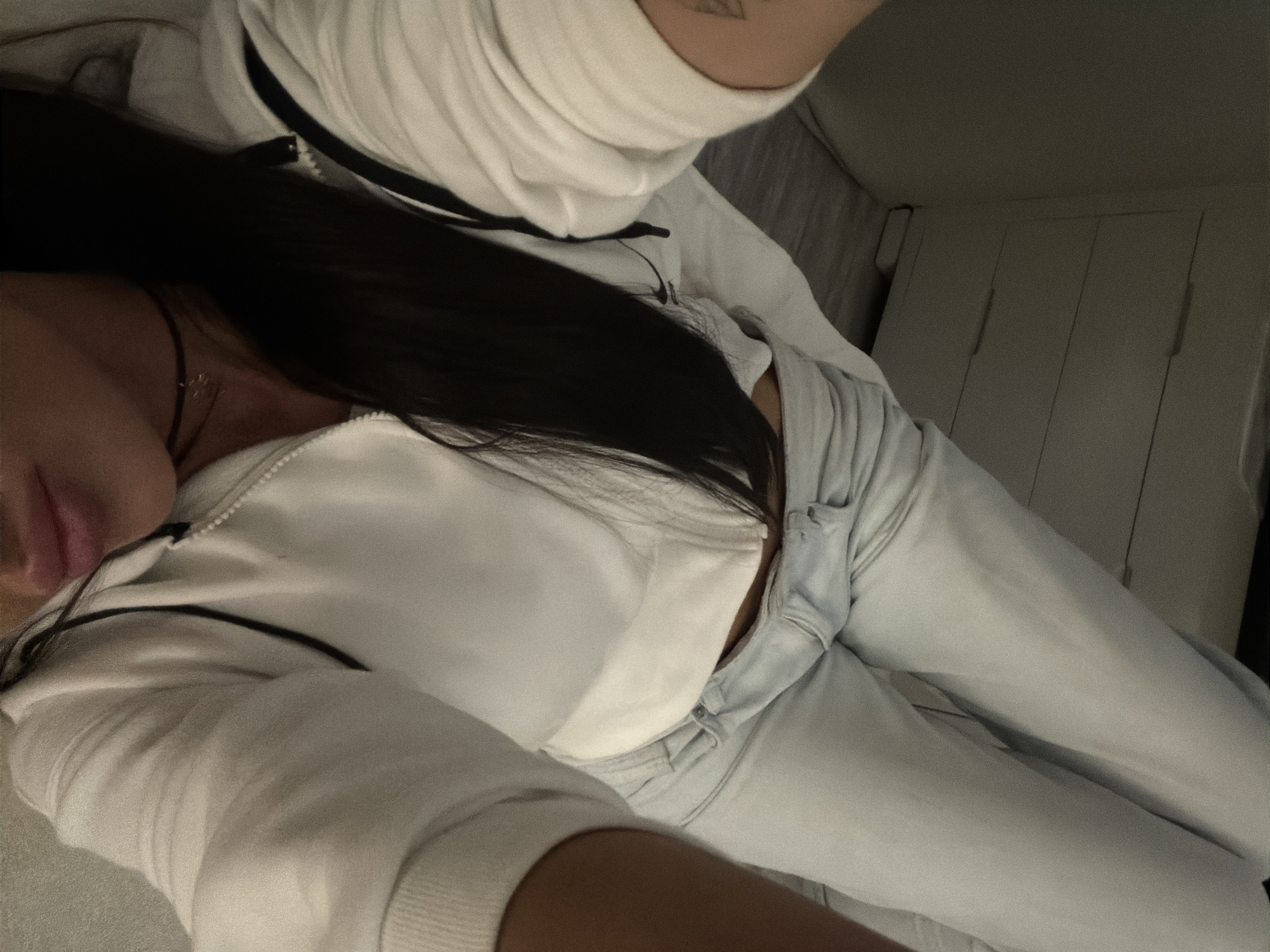 The image shows a person lying on their back, with their legs extended upwards. They are wearing a white hooded sweatshirt and light-colored jeans. Their long, dark hair is draped over their face and chest. The background appears to be a bedroom or living space, with a dresser or cabinet visible on the left and a tiled floor. There is no indication of the location being in Moscow, Russia.
