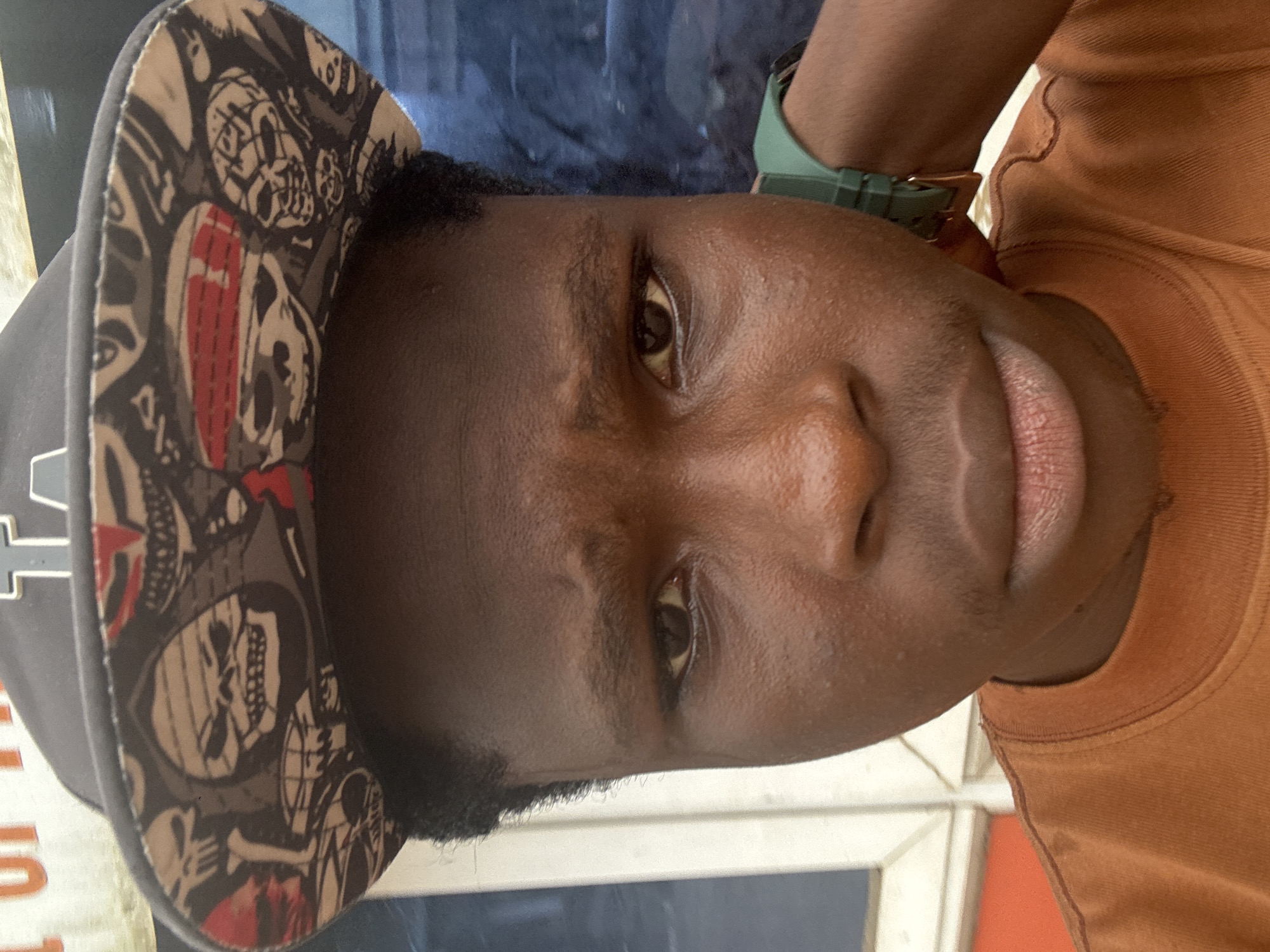 A close-up, head-on, medium shot shows a young Black male wearing a dark baseball cap with a pattern of skulls and a dark orange t-shirt. The cap has a flat brim and appears to be a snapback style. The brim is decorated with a repeating pattern of skulls in various styles, some with red accents. The t-shirt has a ribbed crew neck. The background is out of focus but appears to show some sort of building with blue and white elements, possibly a shop or storefront. A dark green watch is visible on the male's right wrist. The lighting suggests an outdoor setting, possibly during the day. The image is sharp and detailed, highlighting the texture of the subject's skin and the fabric of his clothing. The overall impression is a casual selfie. The location is indicated as Ihiagwa, Nigeria.