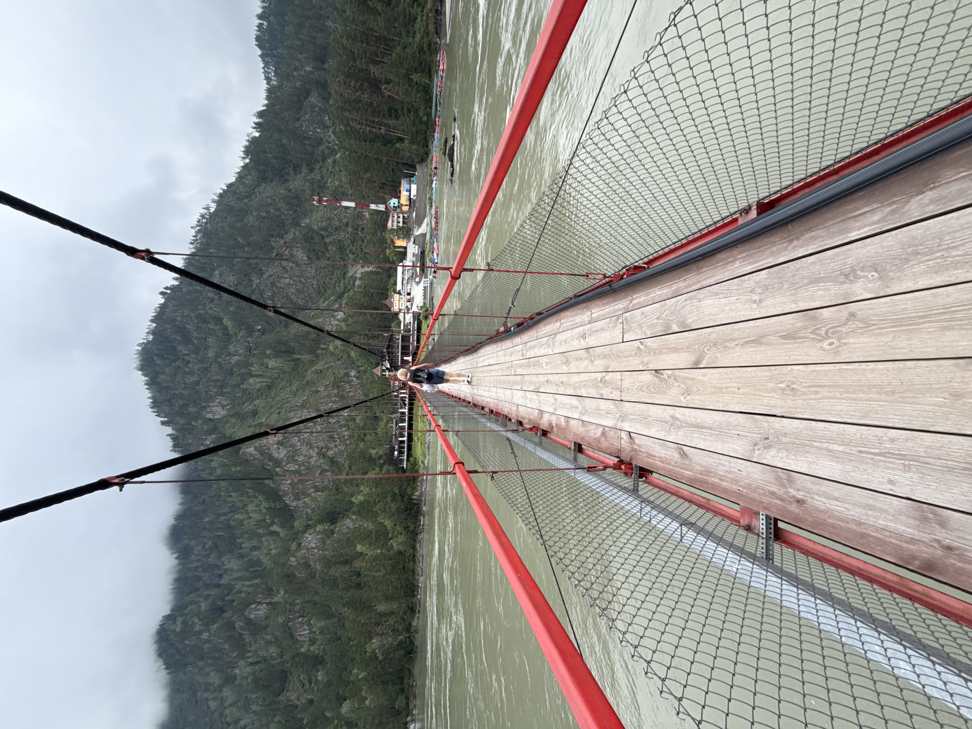 A person wearing a black backpack and blue shorts walks away from the camera on a wooden suspension bridge with red railings and chain-link fencing. The bridge spans a wide, murky green river. In the background, a forested mountain rises under a cloudy sky. On the far bank of the river, there are several buildings, including a red and white tower, and many inflatable rafts are lined up on the shore. The scene is located in Barangol, Russia.