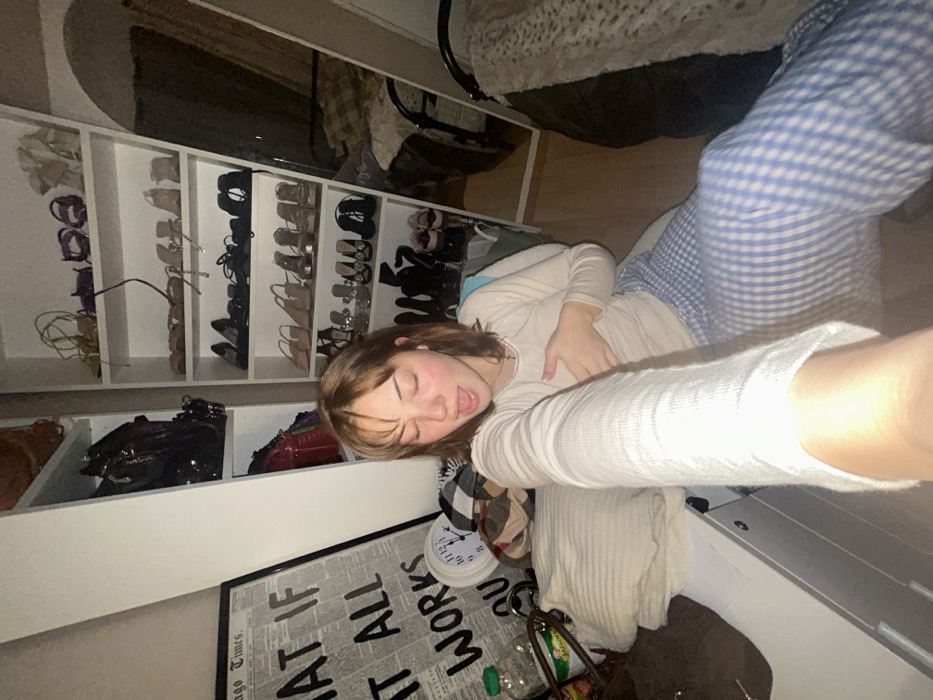 A woman in blue and white pajama pants and a beige sweater lies on the floor, appearing to take a selfie. Behind her, a white shoe rack displays numerous pairs of shoes. A framed newspaper-style poster reading "MAKE IT ALL WORK" sits near a white clock, and scattered items including bags and containers are visible around her. The scene suggests a bedroom or closet space.