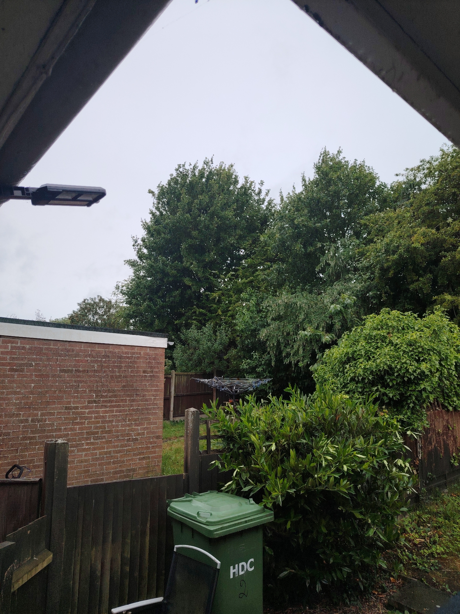 The image displays an outdoor residential scene under an overcast sky, viewed from beneath a dark grey architectural overhang. A modern, dark grey streetlamp fixture is visible in the upper left quadrant.
In the midground on the left, a single-story red brick building with a flat roof and white fascia board is present. To its right, a dark wooden fence with concrete posts separates properties. Behind this fence, a rotary clothesline with garments hangs in the open air, partially obscured by green trees. A variety of dense green trees and bushes occupy the background and midground, creating a thick green canopy.
In the foreground, a dark wooden fence runs horizontally. To the right of the fence, a large green wheelie bin with "HDC" and the number "2" in white text is positioned. A black and white outdoor chair is partially visible in front of and to the left of the bin. Abundant green foliage, including shrubs and bushes, fills the lower right and central foreground. Patches of overgrown grass and dirt are visible near the fences.
The presence of the "HDC" logo on the wheelie bin indicates the location is within the jurisdiction of Huntingdonshire District Council, placing the scene in Huntingdonshire, United Kingdom. No people are visible in the image.