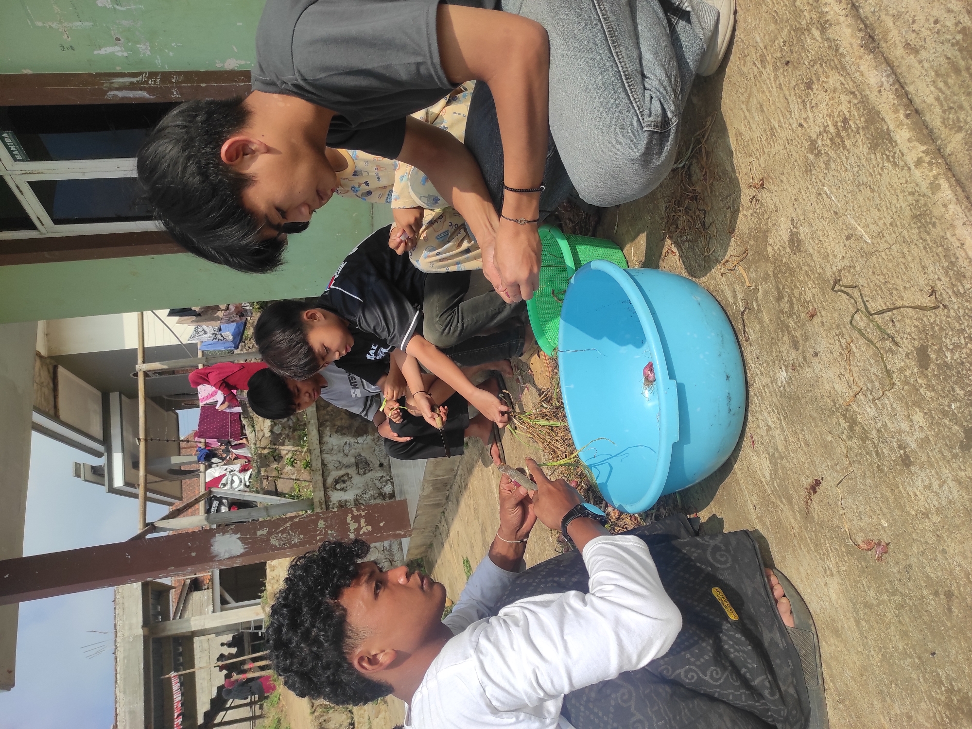 A group of young people are engaged in food preparation outdoors, in what appears to be a residential setting in Sumedang, Indonesia. The central figures, two young men and two younger individuals, are crouched on a concrete ground, focused on processing raw ingredients. One young man, on the left, with curly dark hair, wears a white long-sleeved shirt and a patterned sarong, and is using a knife to trim what look like shallots or spring onions. Beside him, a blue basin contains more of these ingredients, along with a red onion. Another young man, on the right, wearing a dark t-shirt and jeans, is also working with similar produce. The two younger individuals are seated behind them, also participating in the task. In the background, laundry is hanging to dry on a clothesline, and a simple building structure suggests a rural or semi-urban environment. The overall scene conveys a communal activity, possibly related to meal preparation or a farm-to-table practice.