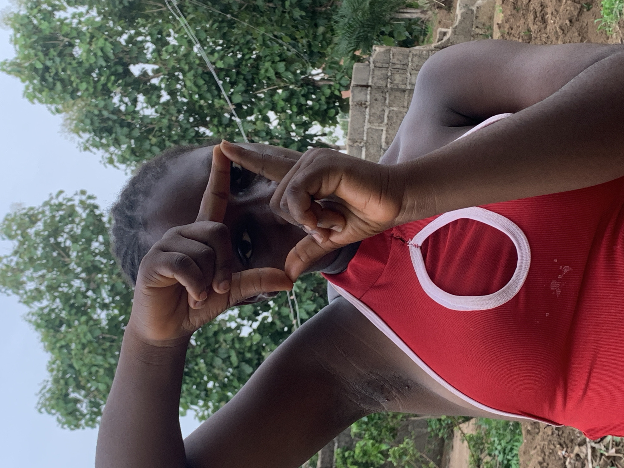 A medium shot shows a person with dark skin and dark hair, wearing a red tank top with white trim, making a rectangular shape with their fingers. The person appears to be outdoors in Agban, Nigeria, as suggested by the presence of green trees and foliage in the background. A brick structure is also visible behind the person. The weather appears cloudy.