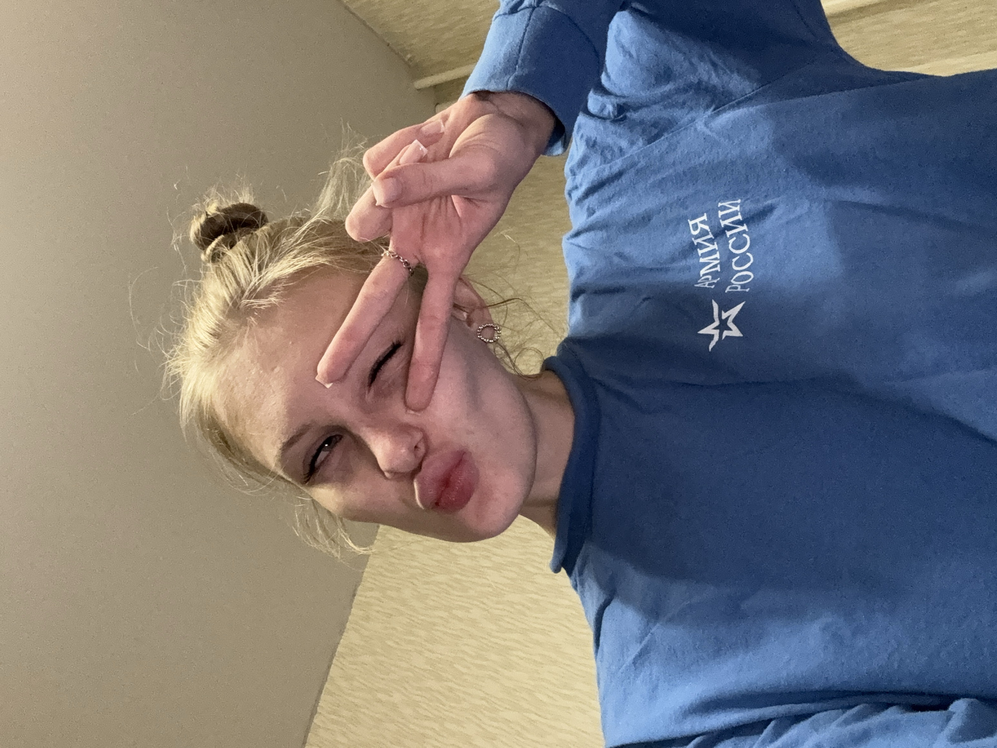 A young woman with blonde hair pulled into a bun is pictured in a selfie. She is wearing a blue long-sleeved shirt with a star emblem and Russian text that reads "АРМИЯ РОССИИ" (ARMY OF RUSSIA). Her right hand is raised, forming a peace sign gesture over her right eye, and she is puckering her lips. She is wearing a ring on her right ring finger and a small hoop earring in her left ear. The background appears to be an indoor wall with textured wallpaper. The lighting suggests it is nighttime or a dimly lit interior. The location is the village of Kondratovo, Perm Krai, Russia.