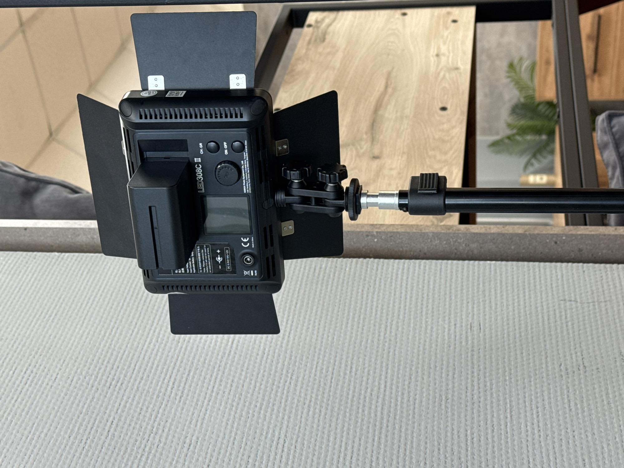 A close-up shot shows a studio LED light (LED308C II) mounted on a black metal boom arm. The light is equipped with black barn doors and a battery pack is attached. The light's control panel, including power and channel/group buttons, is visible. The backdrop includes a white, vertically textured surface, a partially visible gray upholstered sofa, and a glimpse of a wooden-toned structure behind the light, reflecting a green plant. The image does not indicate a location in Chaikovsky, Russia.