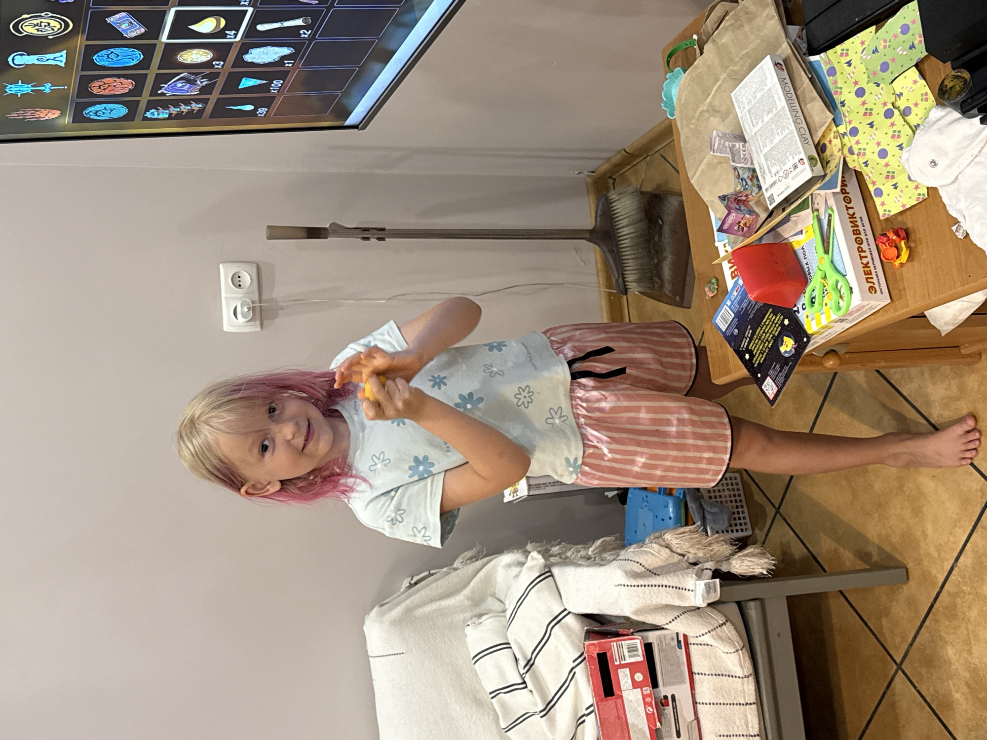 An indoor scene, location Unknown, Unknown, features a fair-skinned child approximately 6-8 years old with shoulder-length blonde hair partially dyed pink, standing and facing the viewer with a slight smile. The child wears a light blue t-shirt patterned with blue flowers and pink and white striped shorts with a black drawstring; the child is barefoot. Both hands grasp and manipulate a small, irregular yellow-orange object.
To the child's left, a light grey upholstered armchair is partially covered by a white blanket featuring black horizontal stripes and tassels. A red and white cardboard box rests on the chair. The wall behind the child is light beige.
To the child's right, a white double electrical outlet with one white power cable plugged in is mounted on the wall. Below the outlet, a brown and grey stand-up broom and dustpan set leans against the wall. Further right, a large flat-screen television displays a video game interface, showing an inventory screen with various item icons, including runes, a book, several vials, a scroll, and quantities (e.g., "x4" next to a yellow drop icon, "x100" next to blue shards, "x39" next to a blue triangle).
In the foreground to the right, a wooden table is laden with multiple objects, including a brown paper bag, a white box with partially legible text such as "MODELING CLAY" (reversed) and Cyrillic text "ЕЛЕКТРОВІКТОРІН," a red rectangular plastic container, green children's scissors, patterned fabric squares (green with yellow trees and dots), a white cloth article with a snap closure, and several small craft items or toys. The floor is covered with light brown square tiles with dark grey grout lines.