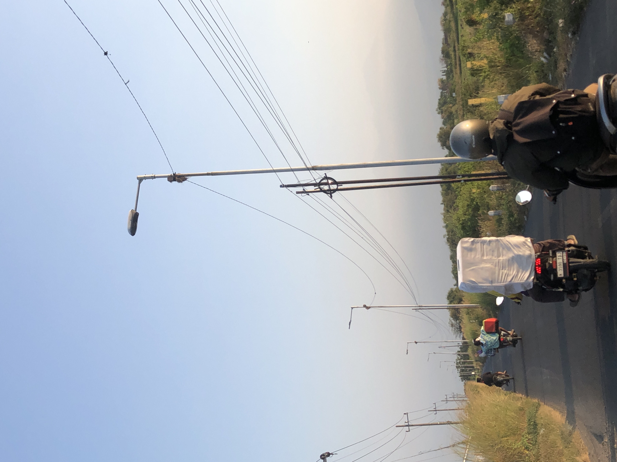 The image shows a paved road with motorcycles driving away from the camera in Glagah Arum, Indonesia. The sky is clear and light blue, with a few wispy clouds in the distance. Several streetlights and utility poles line the side of the road, with power lines stretching across the frame. On the right side of the road, there is a grassy field with some bushes and trees. In the foreground, a person wearing a helmet and a dark jacket is riding a motorcycle, carrying a backpack. Another motorcycle in front of them is carrying a large, white, rectangular object. Further down the road, two more motorcycles are visible, with riders wearing light-colored shirts. The overall impression is one of a rural road in Indonesia during daylight.