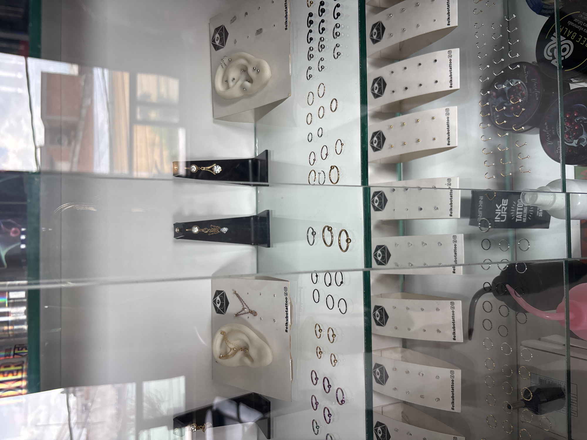 A glass display case in a body piercing and tattoo studio in Cholula, Mexico, presents a diverse collection of body jewelry and related products. The top shelf prominently features two realistic, light-colored ear models positioned on white rectangular display cards. The left ear model showcases an industrial piercing with gold-toned barbells connected by a chain, alongside a helix piercing featuring a gold-toned ring with a chain charm. The right ear model displays multiple helix, conch, and tragus piercings adorned with silver-toned studs and rings. Both display cards are branded with "#elkubotattoo" and include social media icons (Instagram and Facebook). Flanking the ear models are two black stands, each holding a dangling belly button ring; the left one is gold-toned with two clear gems, and the right one is gold-toned with a single clear gem and a decorative butterfly charm.
The middle shelf contains multiple organized trays of individual piercing jewelry. On the left, various seamless rings are displayed in purple, gold-toned, and silver-toned finishes. Adjacent to these are additional trays showcasing gold-toned, silver-toned, and black seamless rings. Further to the right, a section is dedicated to black and silver-toned horseshoe barbells, each with spherical ends. White display cards, similar to those on the top shelf and also branded with "#elkubotattoo," are visible in the background of this shelf.
The bottom shelf features several white display cards, each presenting multiple small stud earrings in gold-toned, silver-toned, or clear gem designs, all marked with "#elkubotattoo" and social media icons. Below these cards are trays containing numerous small, seamless rings in both gold-toned and silver-toned finishes. Towards the center, a black plastic bottle labeled "INK URE TATTOO HEALING 35ml" is visible next to a black squeeze bottle with a pink nozzle. To the right, several circular containers of tattoo aftercare products are displayed, including one with "Vaselma" and "BLACK CHERRY" branding, and another partially visible as "FREE BALM". The far right section of the bottom shelf holds trays of hook-style nose rings, available in gold-toned and silver-toned finishes, each featuring a small clear gem. Reflections on the glass case reveal portions of an exterior building with brick walls and white structures, indicating an urban setting.