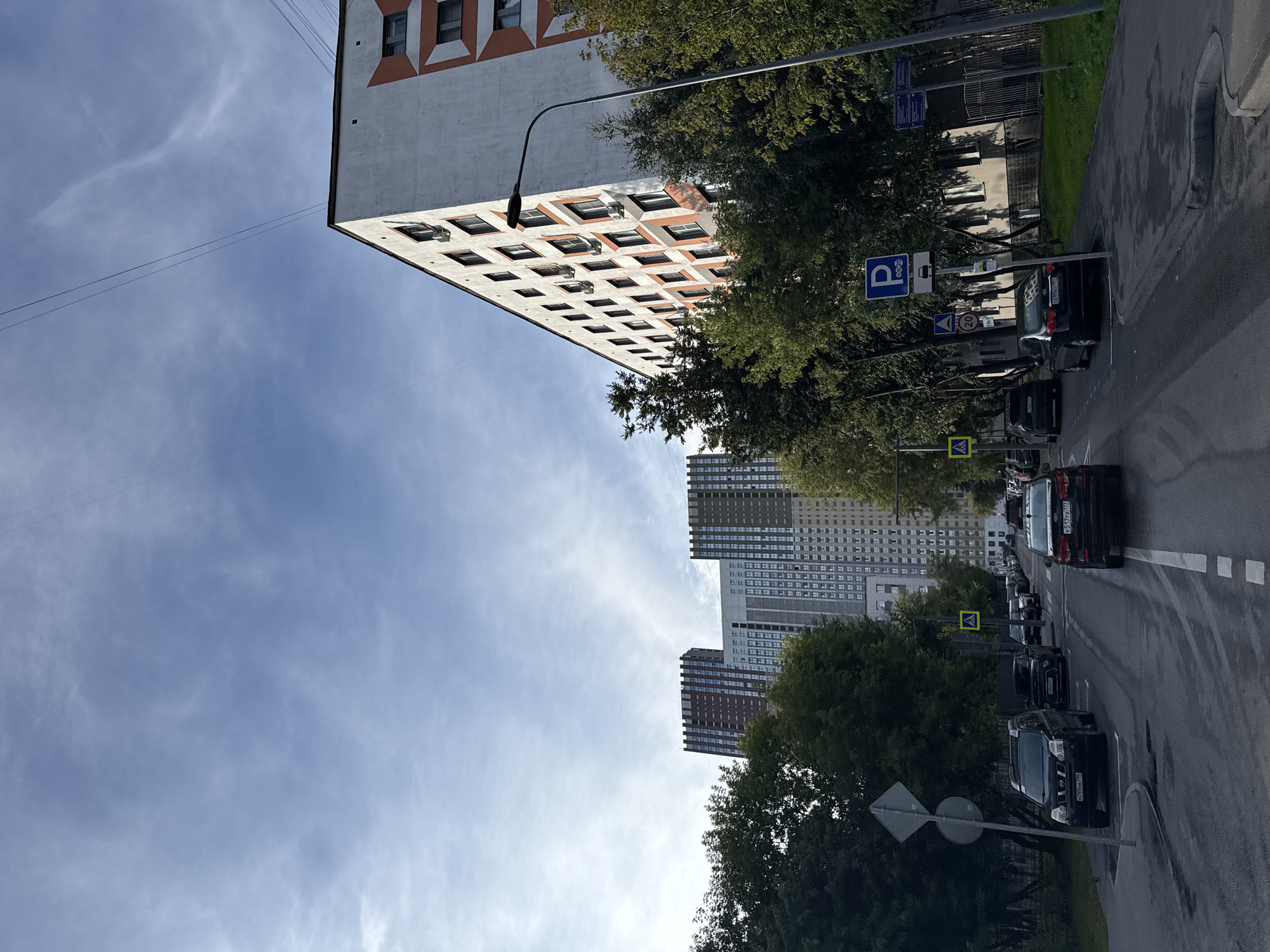The image depicts a street view in Moscow, Russia, on a partly cloudy day. The foreground shows a wet asphalt road with several cars parked and driving. The perspective is from a vehicle moving along the road. Tall apartment buildings, characteristic of Soviet-era architecture, line the street, with one prominent white and orange building on the right and other multi-story buildings visible in the distance. Trees with green foliage line the sidewalks, indicating it is likely late summer or early autumn. Road signs, including parking signs and a pedestrian crossing sign, are visible. The sky is a pale blue with wispy white clouds and a few power lines stretching across it.