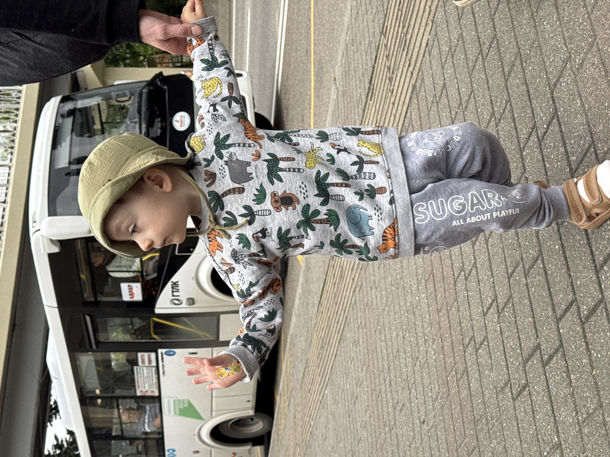 A young child in a tan hat, wearing a gray animal-print sweater and gray pants with "SUGAR" printed on the leg, stands on a paved walkway. The child is holding something small and colorful in their outstretched hand. The child is being held by an adult. A white and black bus is visible in the background. The bus has the word "TTK" in Cyrillic script on its side. The location is most likely a street in the city of Sochi, Russia, as indicated by the Cyrillic script on the bus.