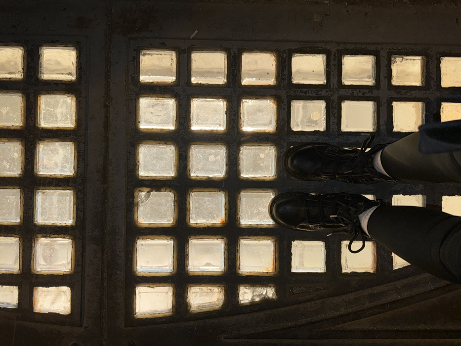 A high-angle photograph shows the lower legs and feet of an individual positioned atop a floor composed of numerous square, translucent glass blocks set within a dark, robust framework. The glass blocks are illuminated from below, emitting a bright, diffuse light. The individual wears black, lace-up combat boots with buckle elements, dark trousers, and white socks. The dark framework exhibits minor surface abrasion and occasional reddish-brown discoloration, likely oxidation, at the periphery of some glass inserts. This distinctive illuminated glass floor is a characteristic architectural element of the Underground Museum within Zaryadye Park, Moscow, Russia.