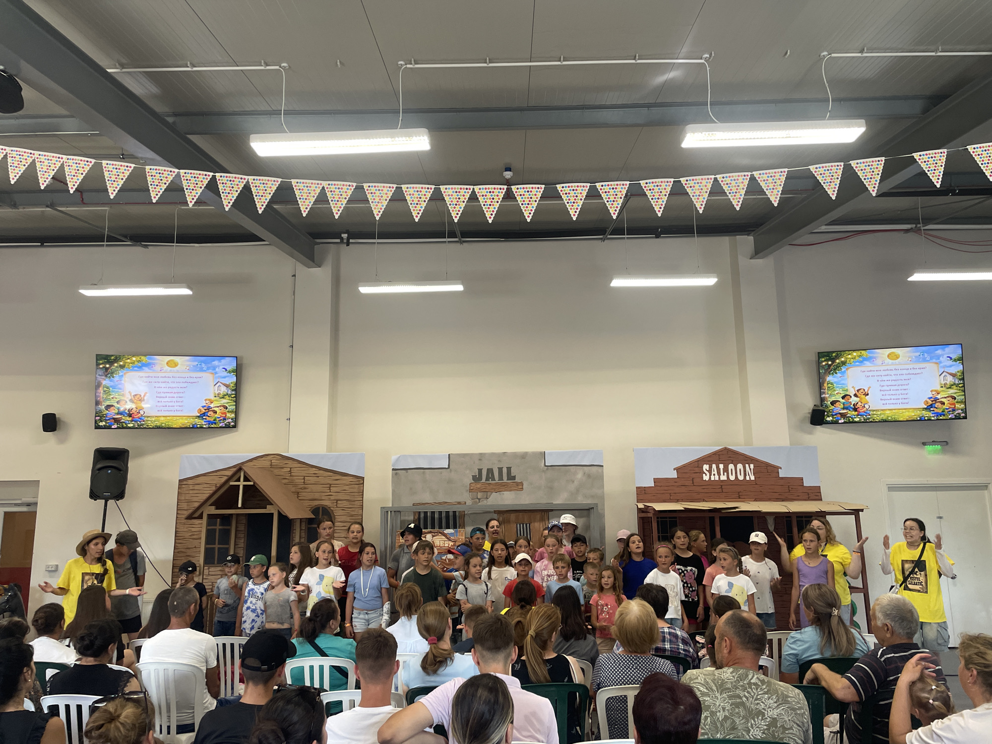 In Zîrnești, Moldova, a group of children are performing on a stage set up in an indoor venue, likely an event hall or community center. The stage features a backdrop with three distinct sections: a wooden house on the left, a building labeled "JAIL" in the center, and a building labeled "SALOON" on the right, suggesting a Wild West theme. A string of colorful triangular pennant flags hangs across the ceiling above the stage.
A group of children, varying in age and gender, are standing in rows on the stage, appearing to sing or act. Two adults are also on stage, one in a yellow shirt to the left, and another in a yellow shirt to the right, both gesturing as if directing or participating in the performance. In the background, a large screen displays an animated image with text in a foreign language, possibly illustrating a story.
The audience, seated in rows of chairs, consists of adults and some children. The environment appears to be a functional indoor space with exposed ceiling structures and fluorescent lighting. The overall scene captures a community event or a school performance.