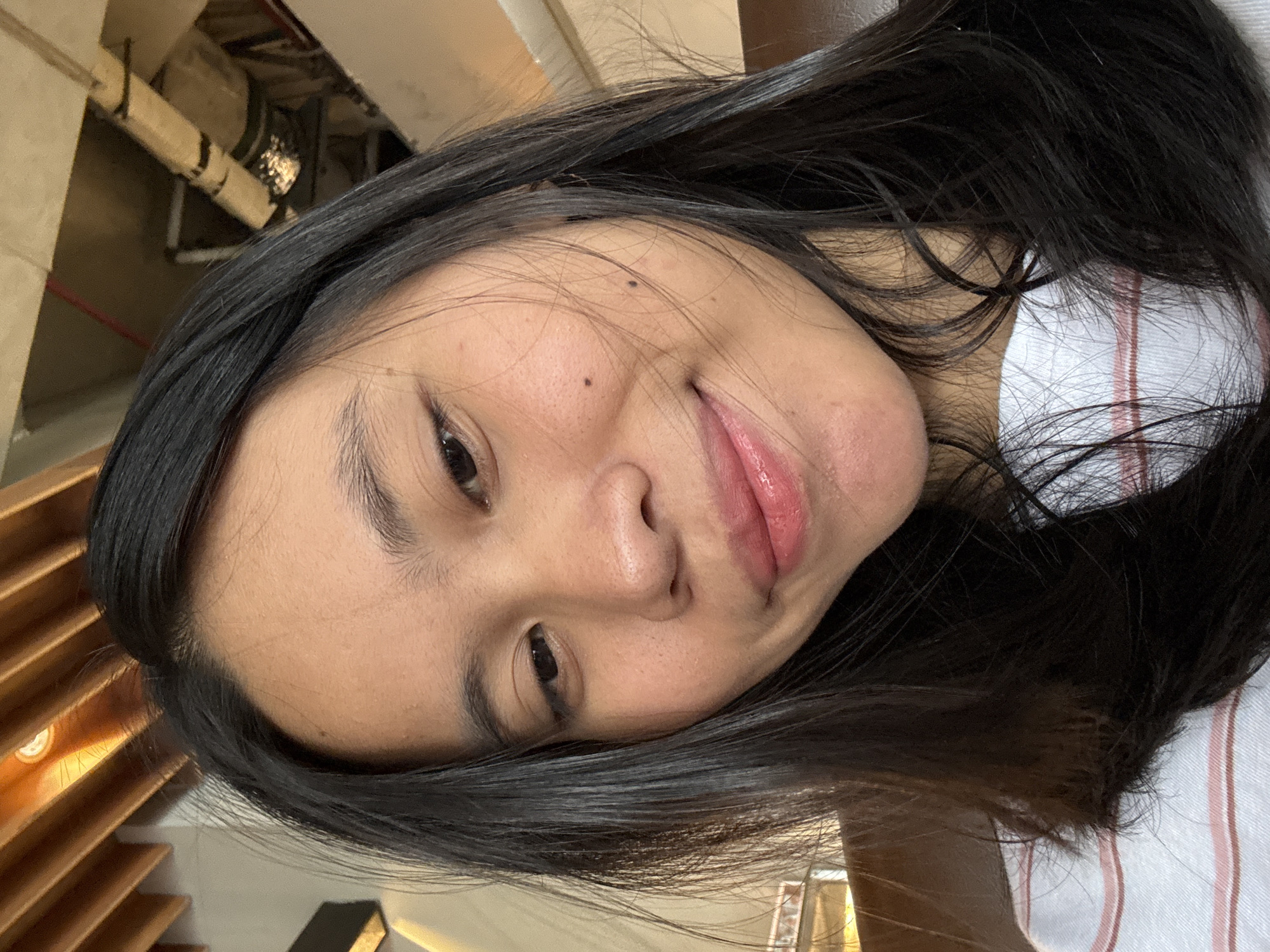 A young woman with long, dark hair and a light complexion is captured in a close-up selfie. She is wearing a white t-shirt with thin pink and blue stripes. Her eyes are looking down and slightly to her left, and she has a subtle smile on her lips, which are enhanced with a rosy lipstick. A small mole is visible on her right cheek. The background appears to be indoors, with wooden beams and industrial ceiling elements visible above and behind her. This photograph was taken in East Jakarta, Indonesia.